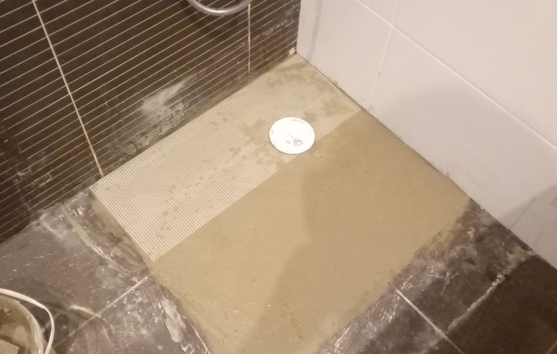 Piso do chuveiro com cimento fresco ao redor do ralo. Os azulejos estão parcialmente cobertos e alguns estão faltando.