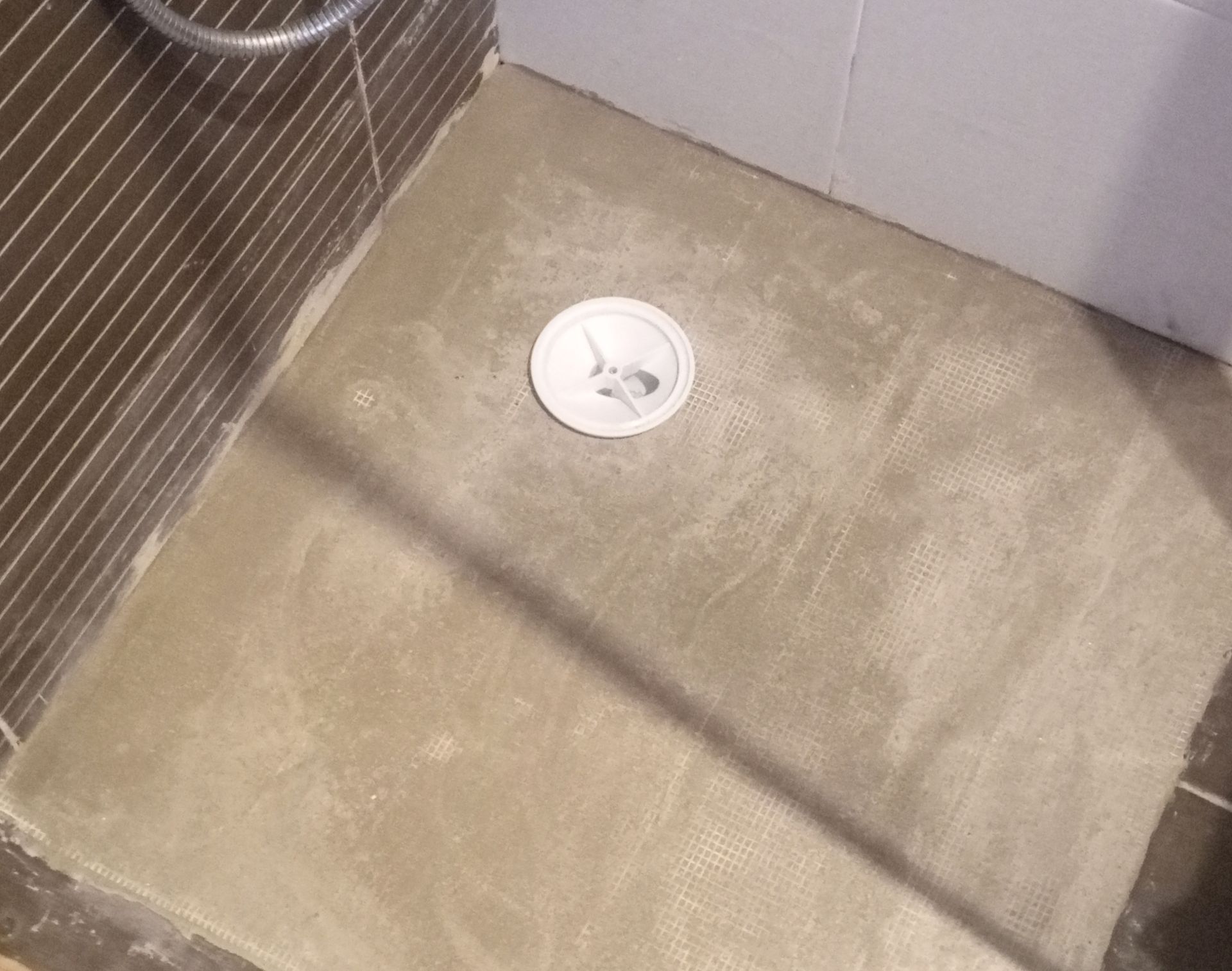 Chuveiro com piso e ralo. Superfície de concreto cinza, tampa de ralo branca, paredes revestidas com azulejos marrom-escuros e brancos.