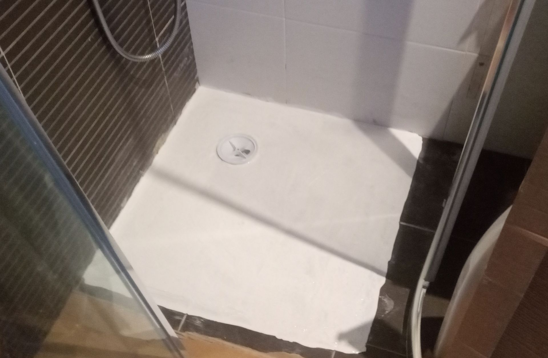 Cabina de duche branca com ralo, parede revestida a azulejos castanhos e porta de vidro.