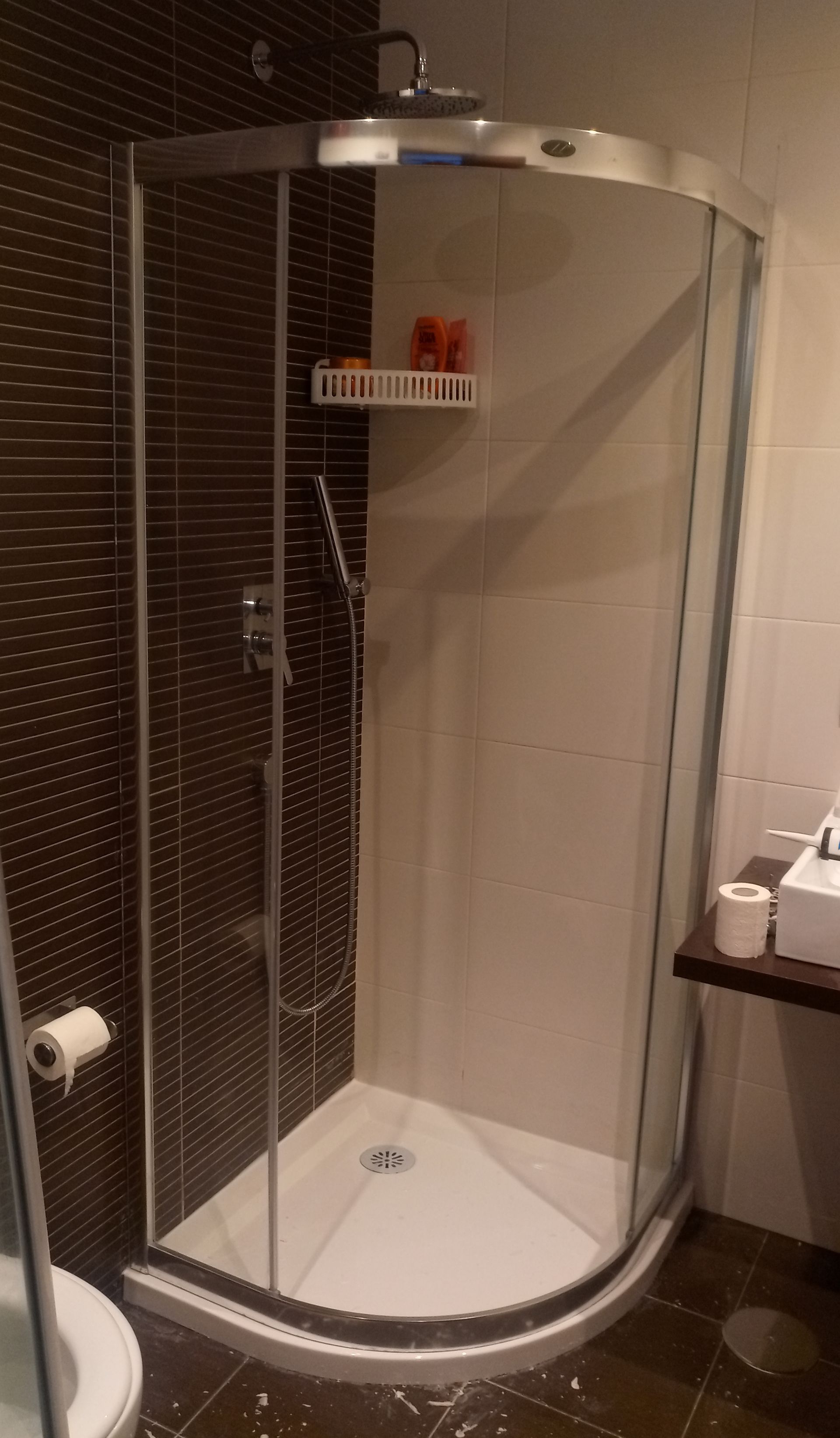 Box de chuveiro com vidro curvo, interior branco, parede revestida com azulejos marrons e prateleira embutida com produtos de higiene pessoal.