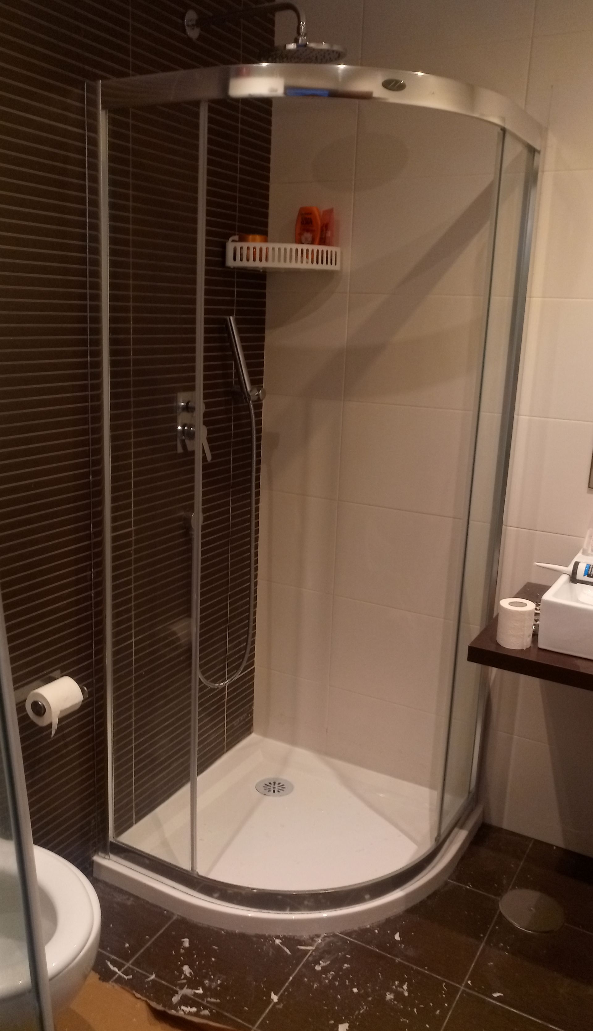 Box de canto para chuveiro com portas de vidro, paredes brancas e fundo de azulejos marrom-escuro.