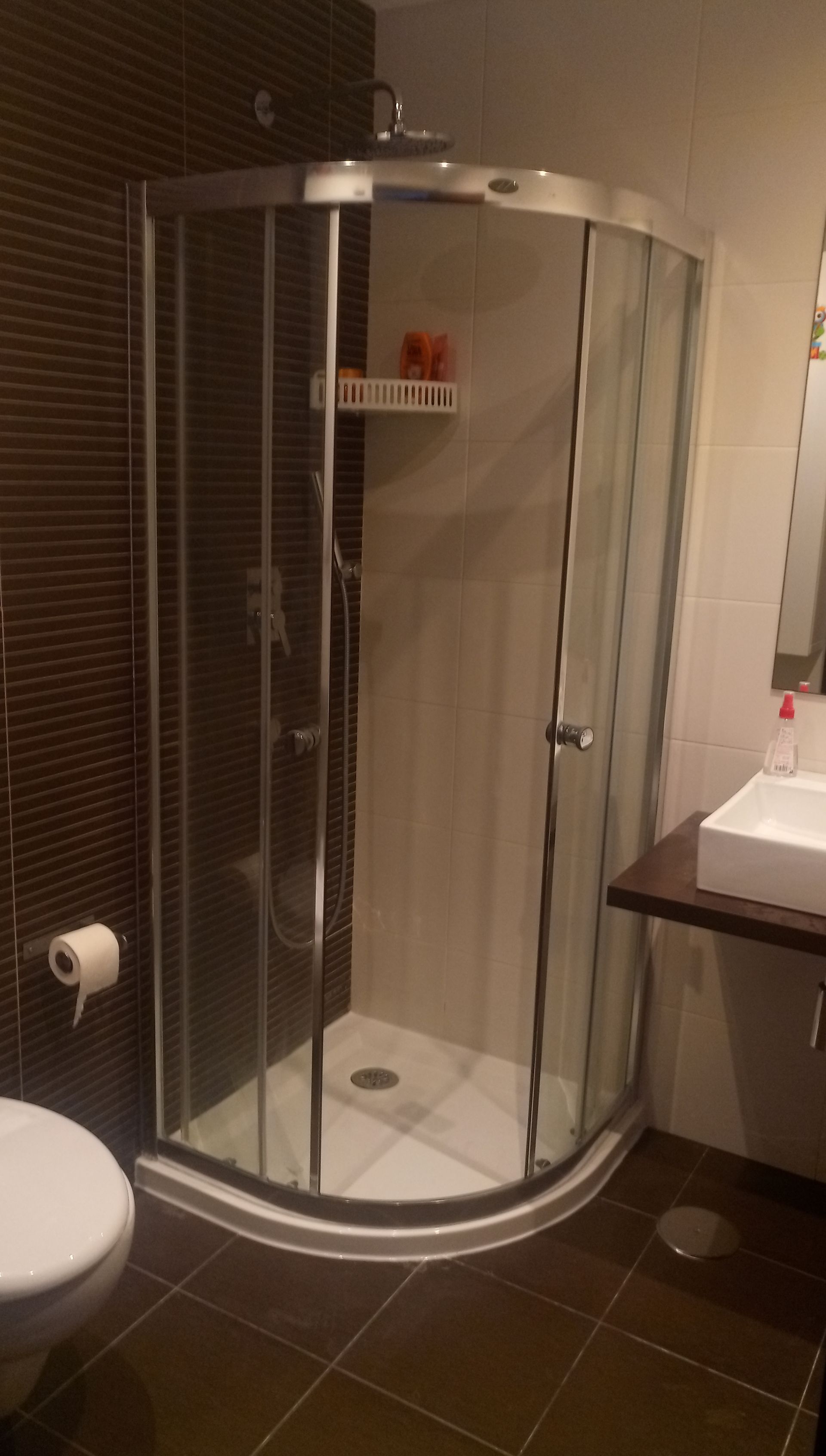 Box de canto com portas de vidro, vaso sanitário à esquerda, pia à direita. Azulejos marrons, detalhes em branco.