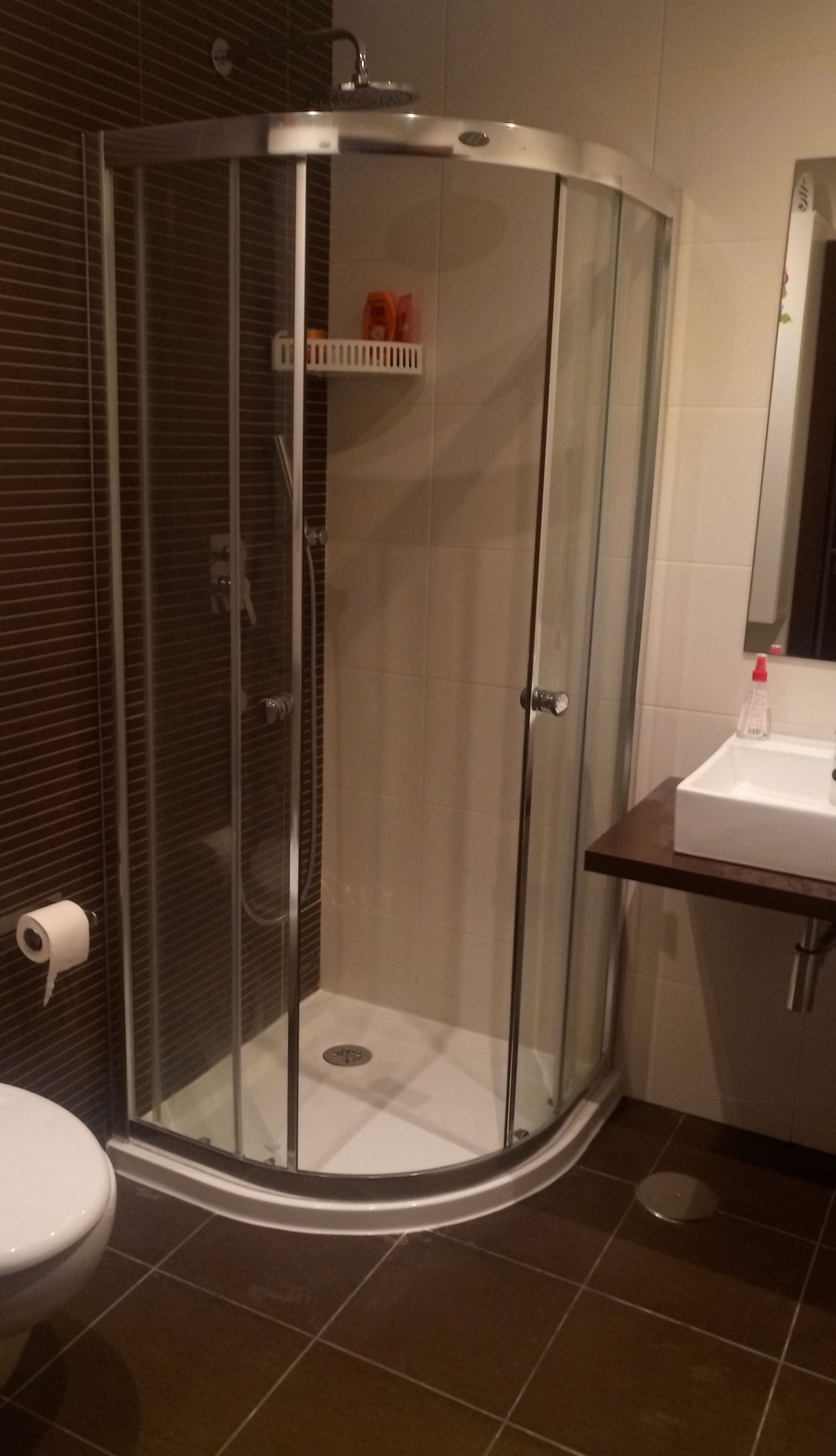 Um chuveiro com porta de vidro curva em um banheiro com azulejos marrom-escuros.