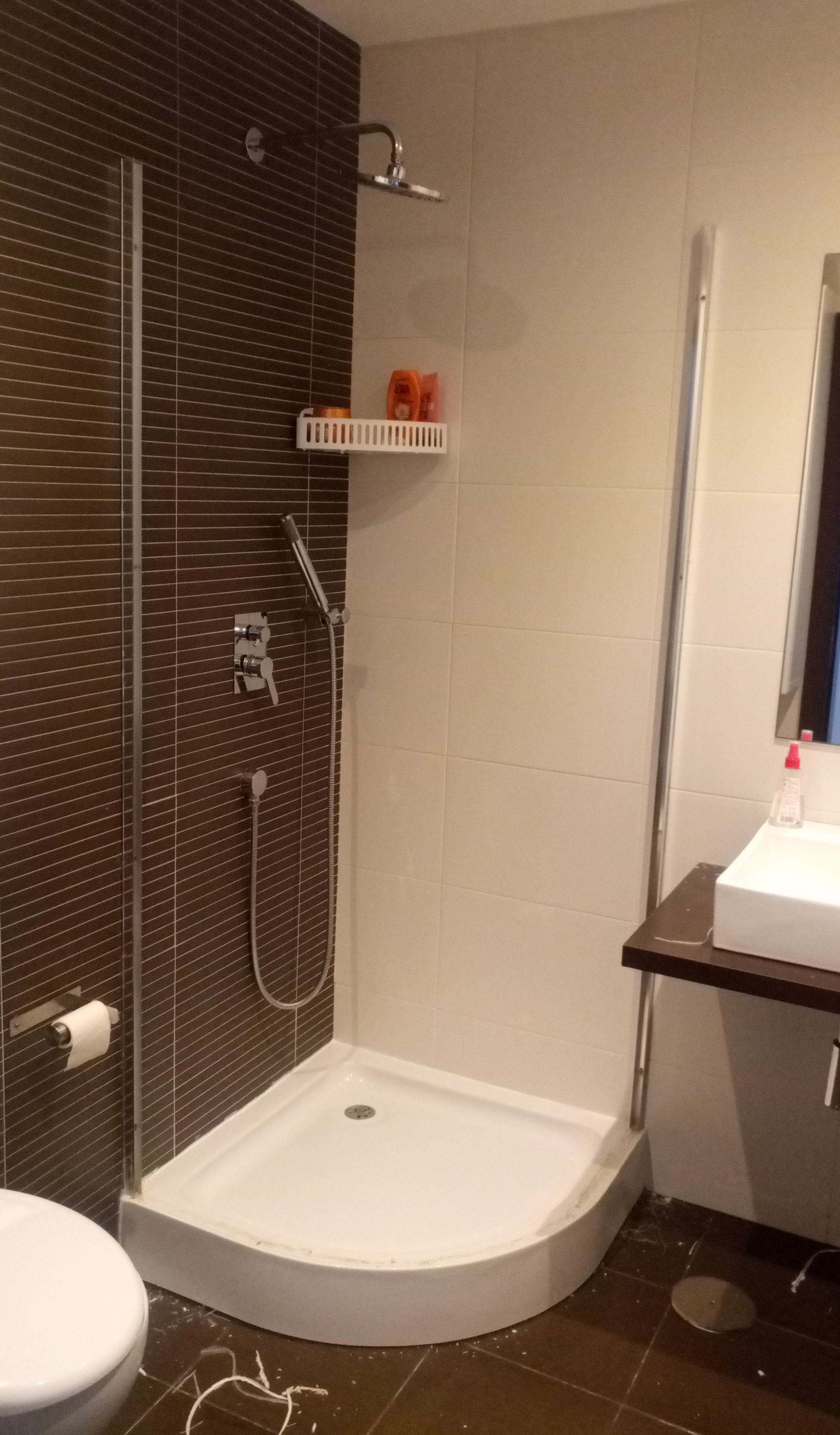 Cabine de duche com base curva branca, parede revestida a azulejos castanhos, chuveiro de mão e prateleira para sabonete.