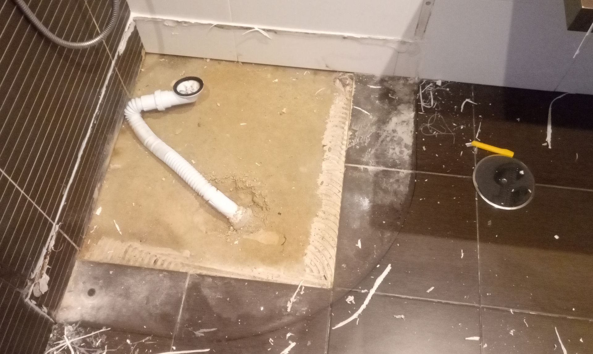 Componentes de drenagem de chuveiro sobre base de concreto, piso de azulejo parcialmente instalado, com tubo de drenagem branco.