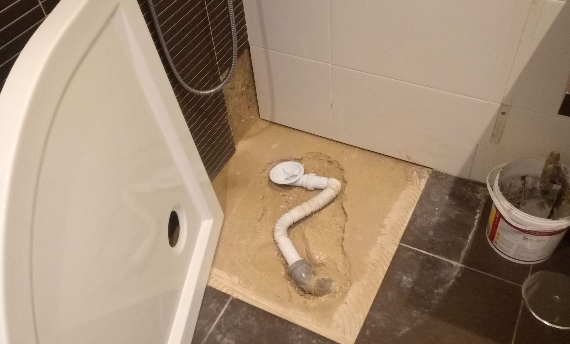 Box de chuveiro em construção, ralo exposto, piso inacabado, balde de materiais.