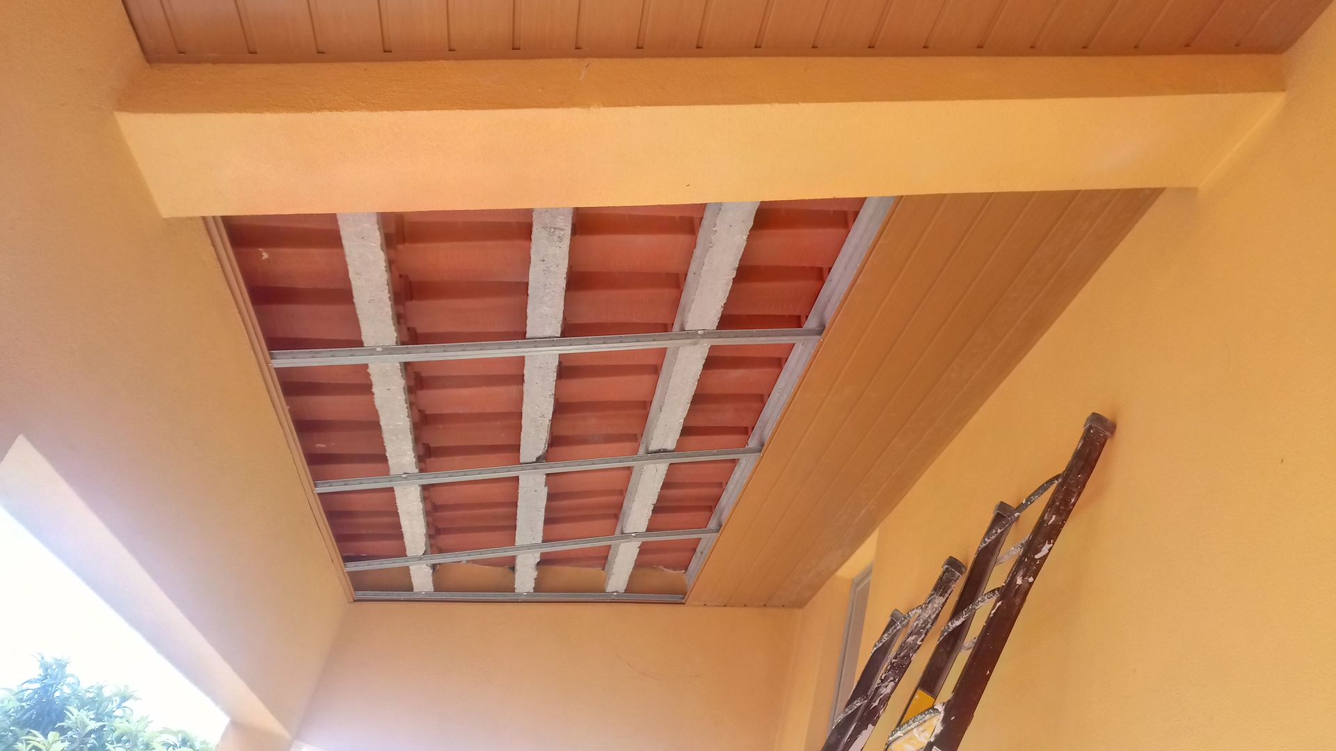 Teto de estuque laranja com telhas vermelhas expostas e vigas de metal. Uma escada está encostada na parede.