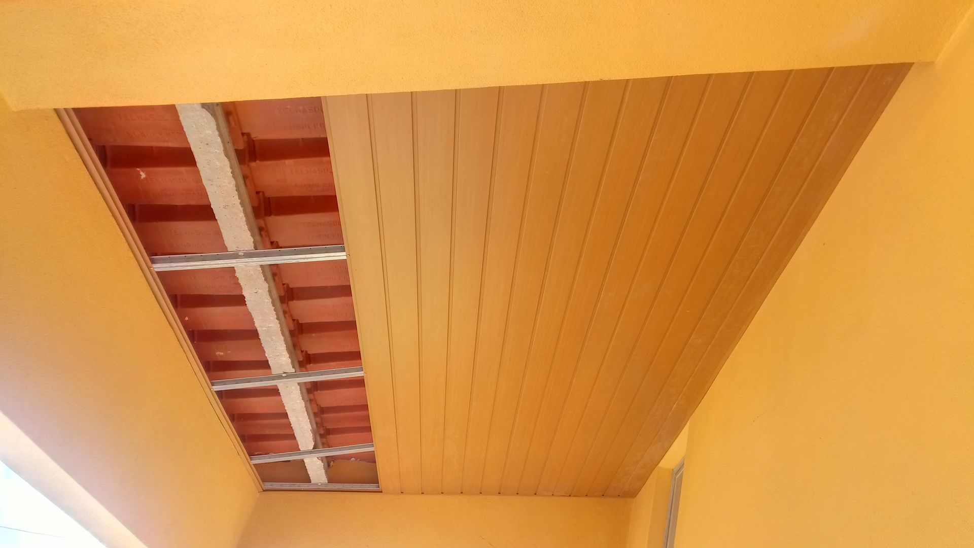 Teto pintado de laranja com painéis de madeira à direita e telhas vermelhas expostas com suportes metálicos à esquerda.