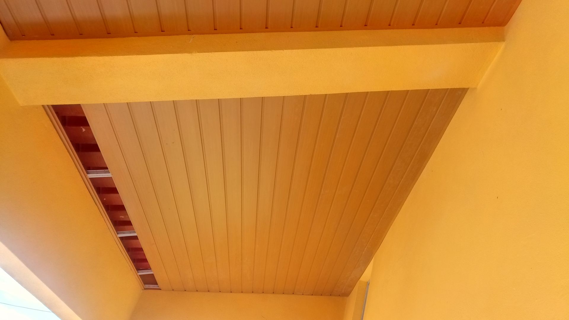 Teto pintado de amarelo com tábuas de madeira, vigas e uma área exposta com azulejos vermelhos.