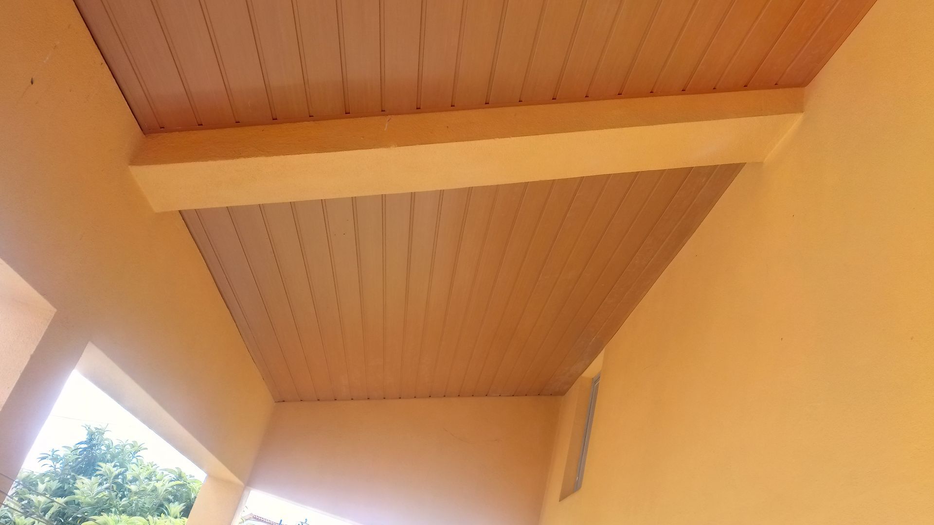 Teto pintado de bege com tábuas de madeira, sustentado por uma viga cor creme, contra paredes amarelas.