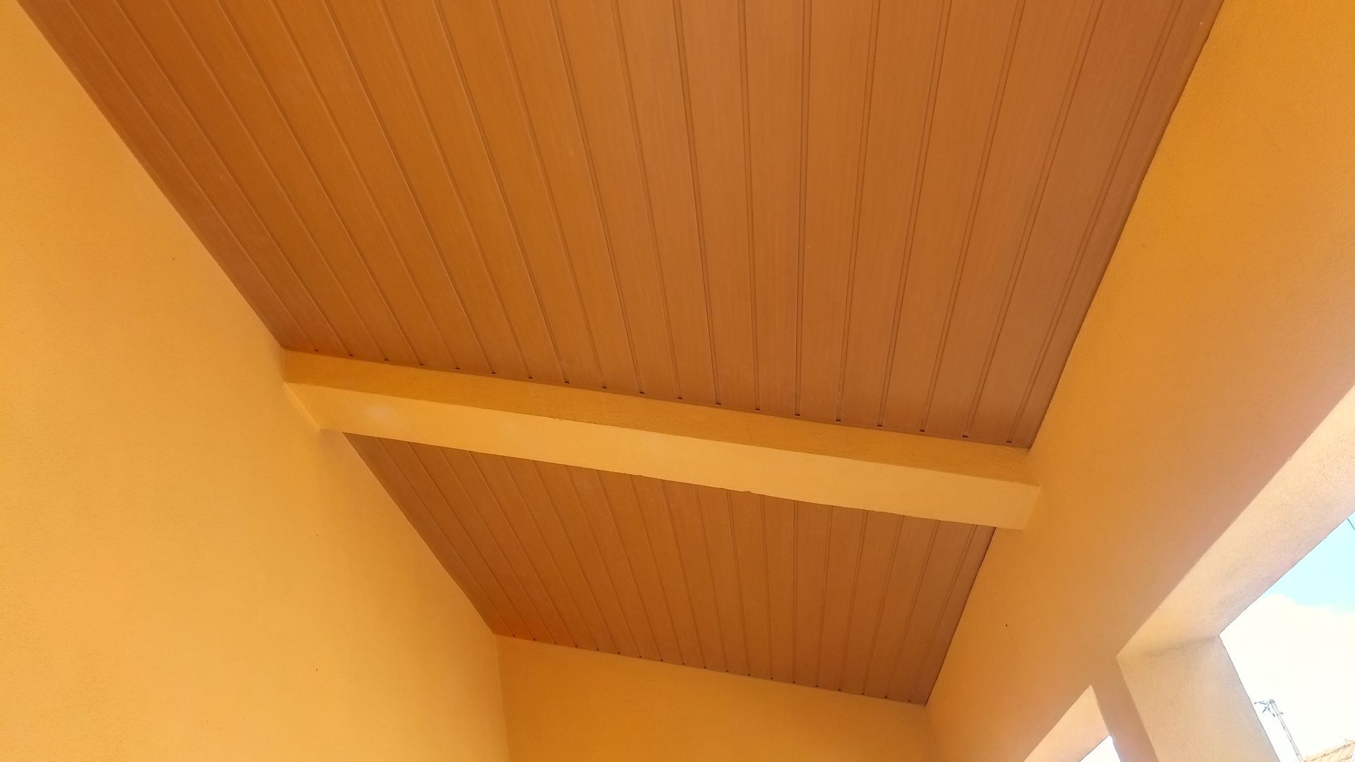 Teto de madeira pintada de bege com viga de sustentação, cercado por paredes bege.