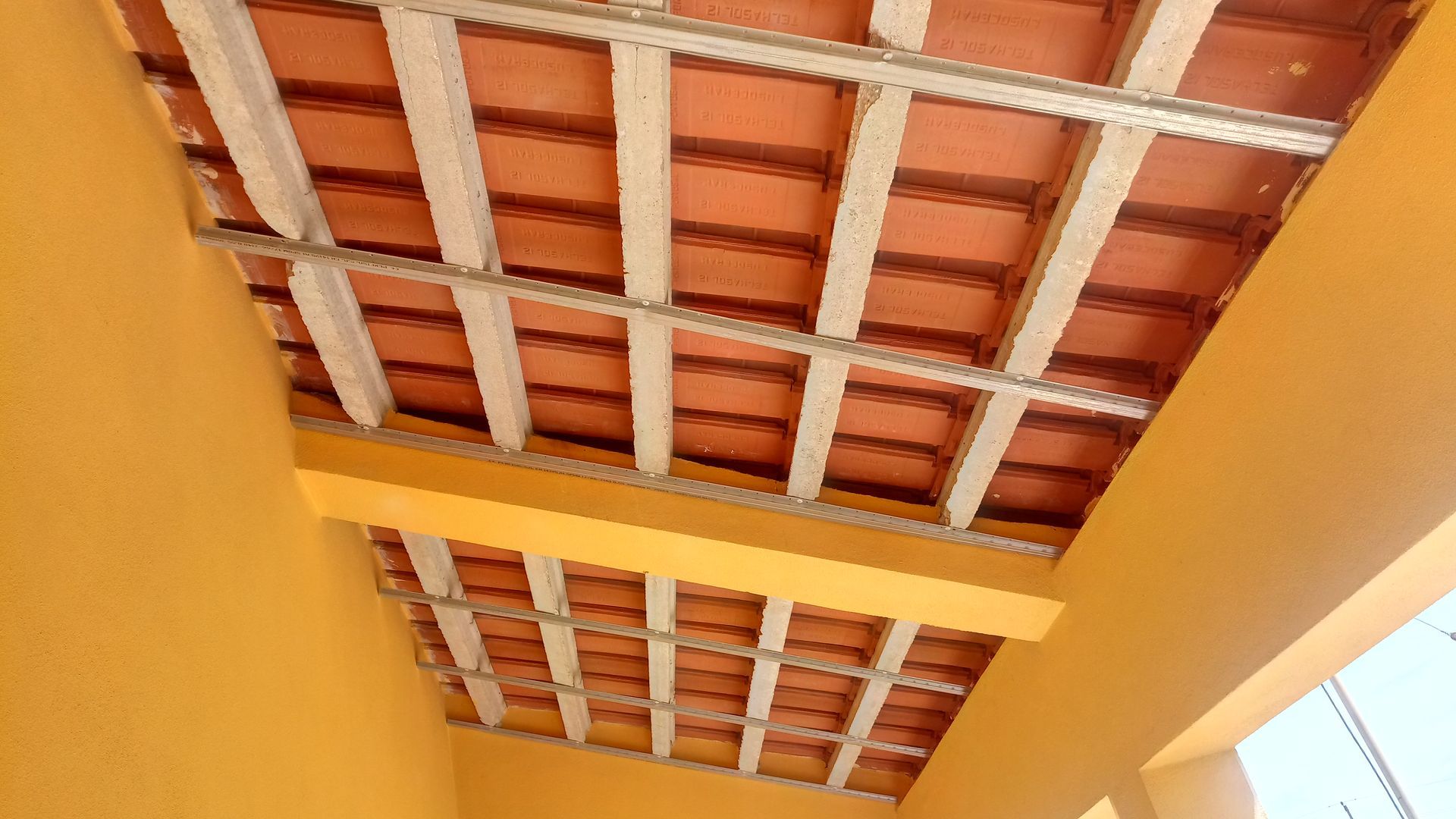 Vista de um teto de telhas de terracota sustentado por vigas de metal, situado em um edifício pintado de amarelo.