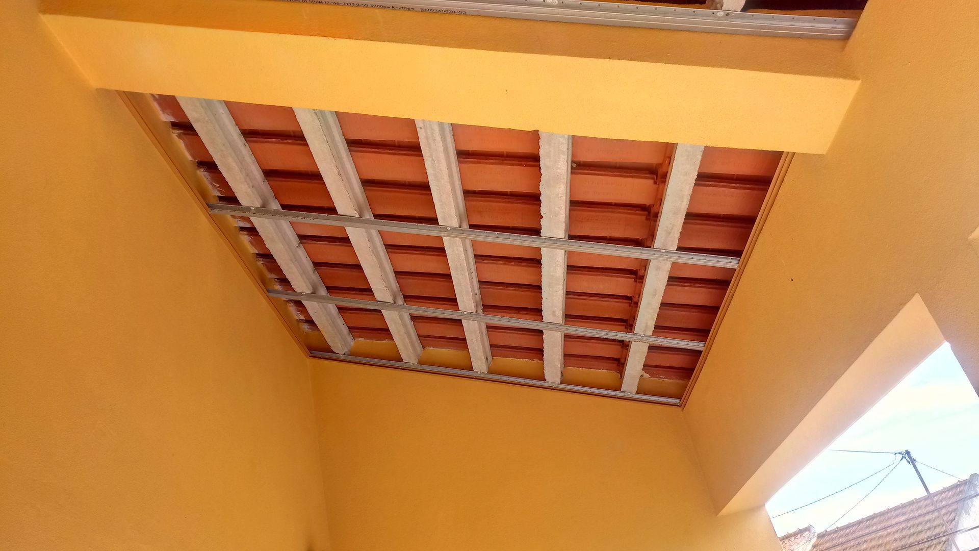 Teto pintado de laranja com telhas vermelhas expostas, sustentado por vigas metálicas.
