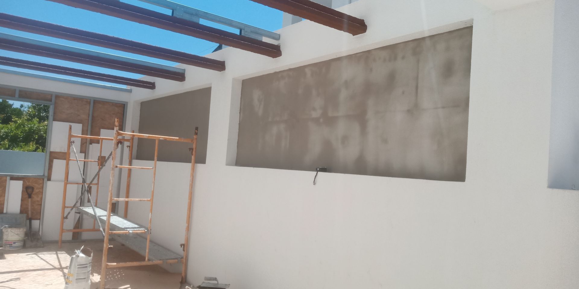 Canteiro de obras com paredes brancas, vigas expostas, andaimes e janelas cobertas. Céu azul visível.