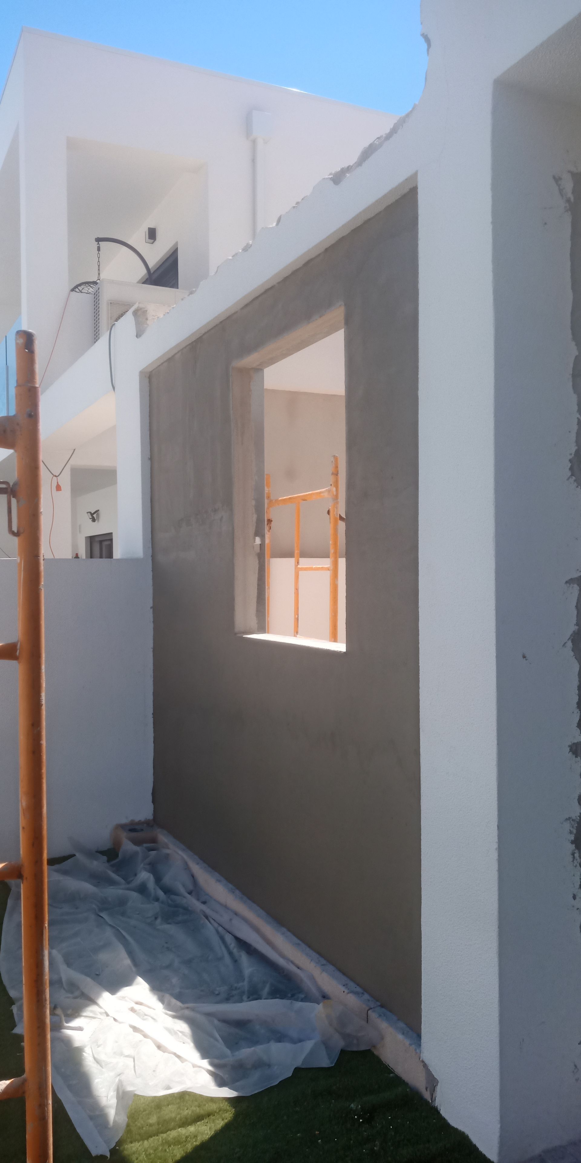 Parede externa de um edifício em construção, apresentando uma janela e cimento aplicado contra uma estrutura branca.