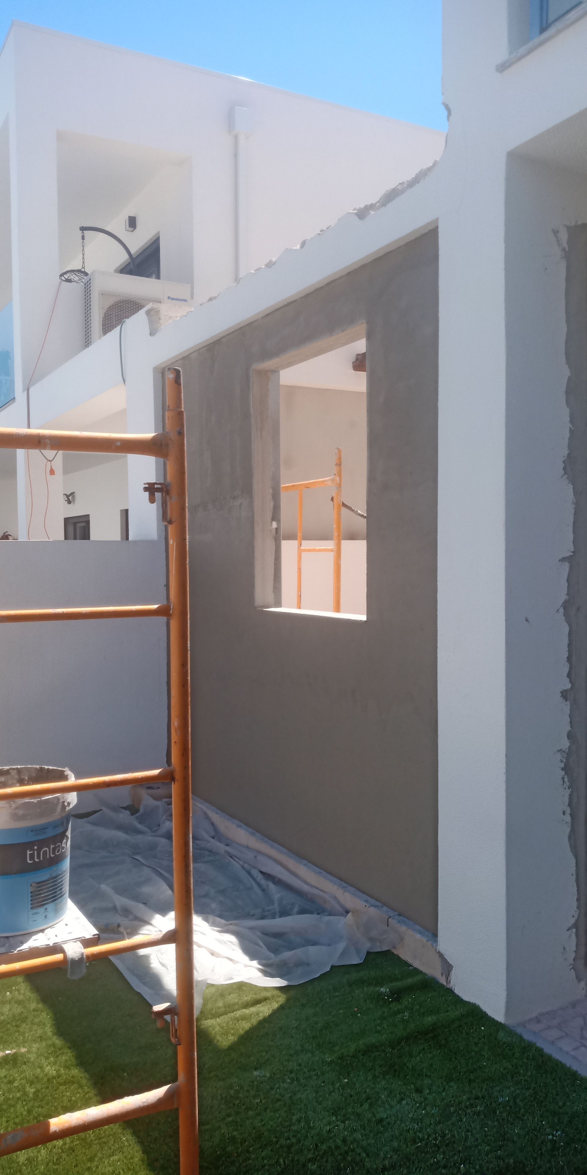 Parede externa em construção com abertura para janela, andaimes e grama.