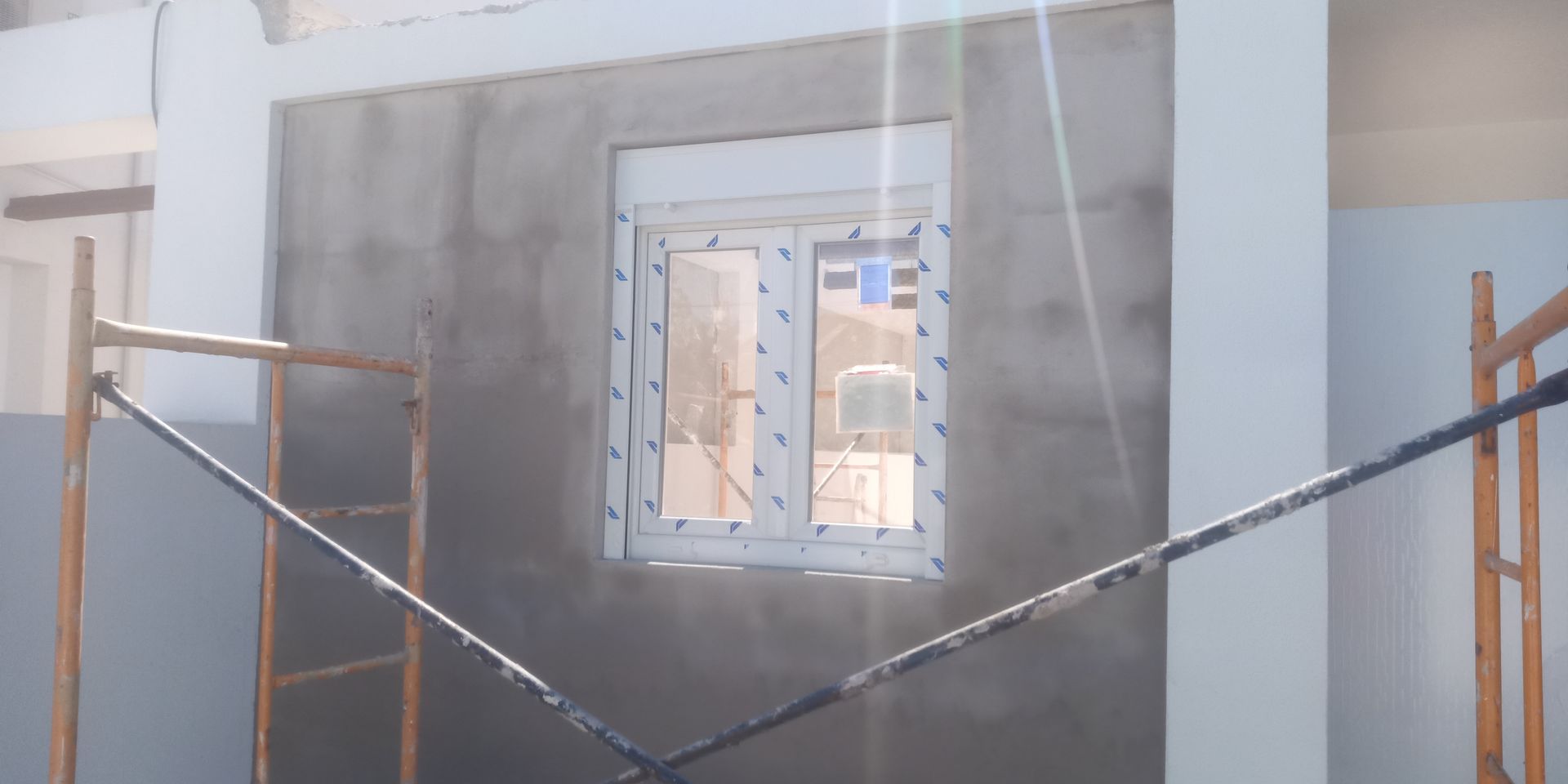 Parede externa com janela, cercada por andaimes. A parede é revestida com gesso cinza.