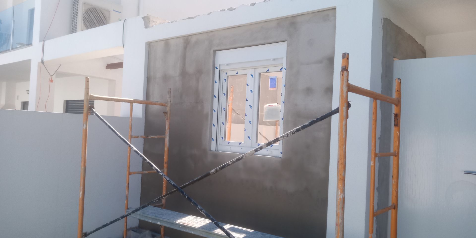 Vista exterior de um edifício em construção. Uma janela está inserida numa parede rebocada a cinzento. É possível ver os andaimes.