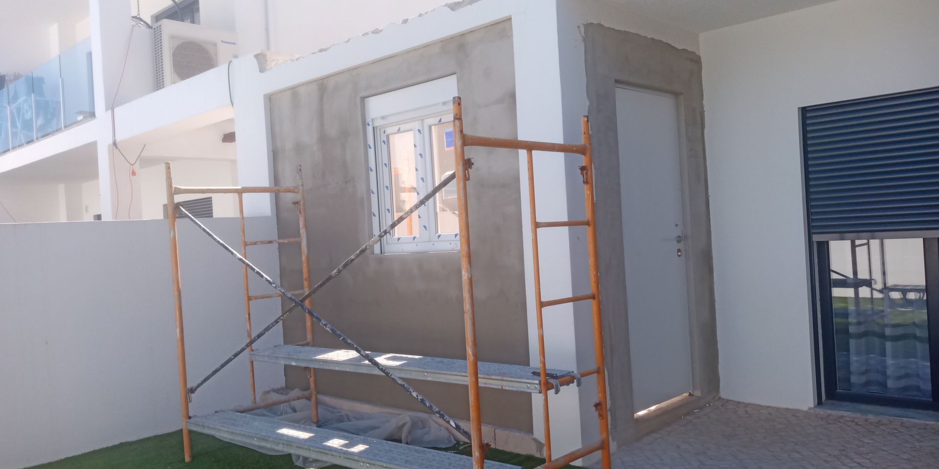 Canteiro de obras com andaimes. Janela e porta emolduradas em uma parede de estuque cinza.