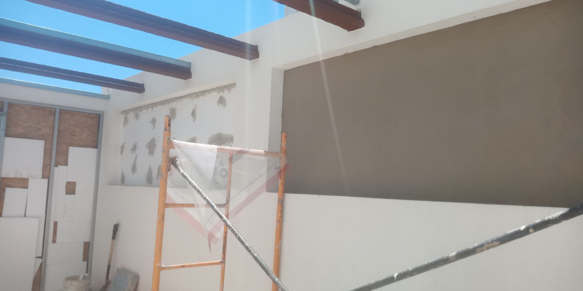 Canteiro de obras com paredes brancas, escada, vigas e uma grande janela.