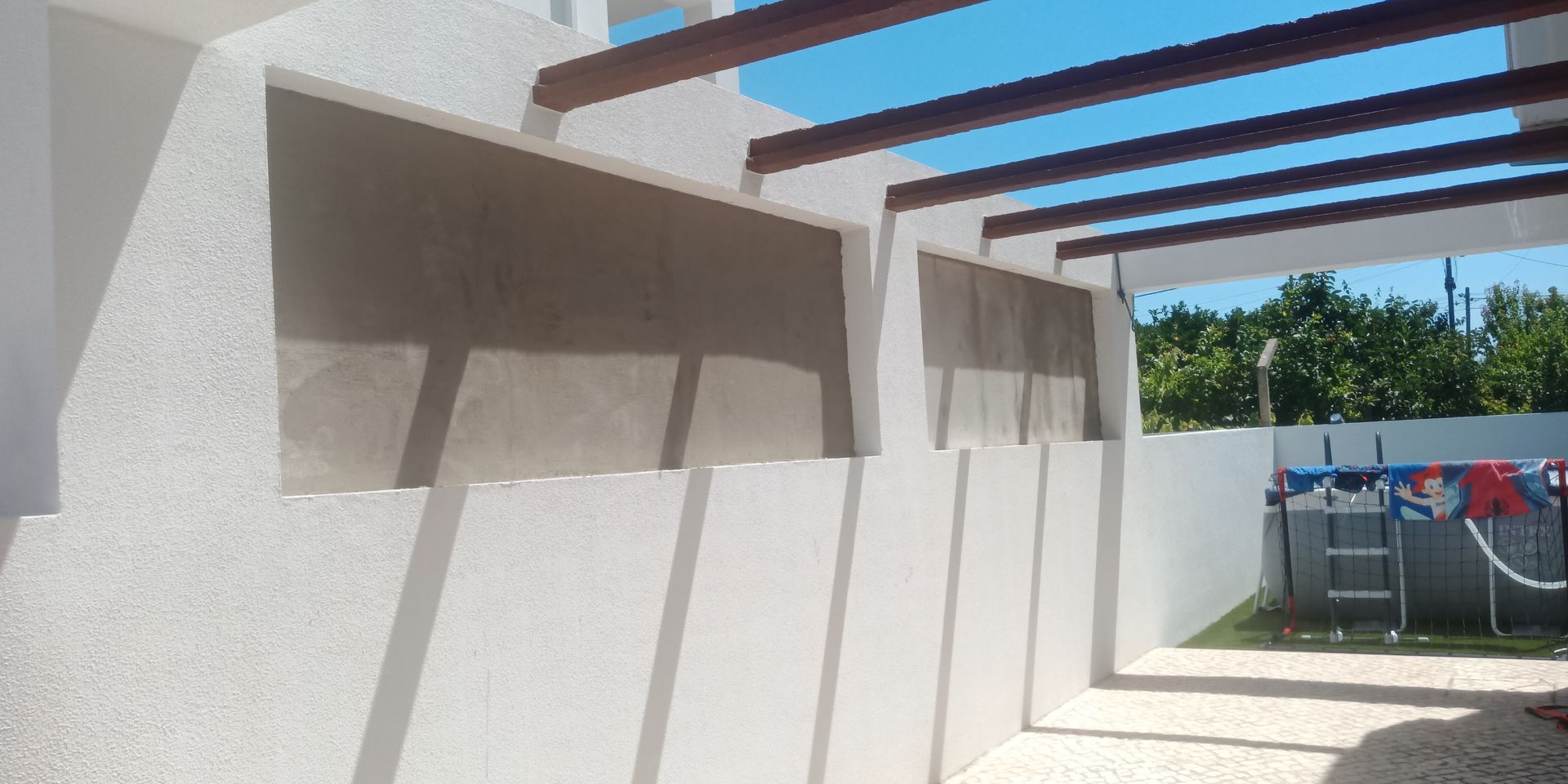 Parede de estuque branco com aberturas retangulares, vigas de madeira no teto, vista para o quintal.