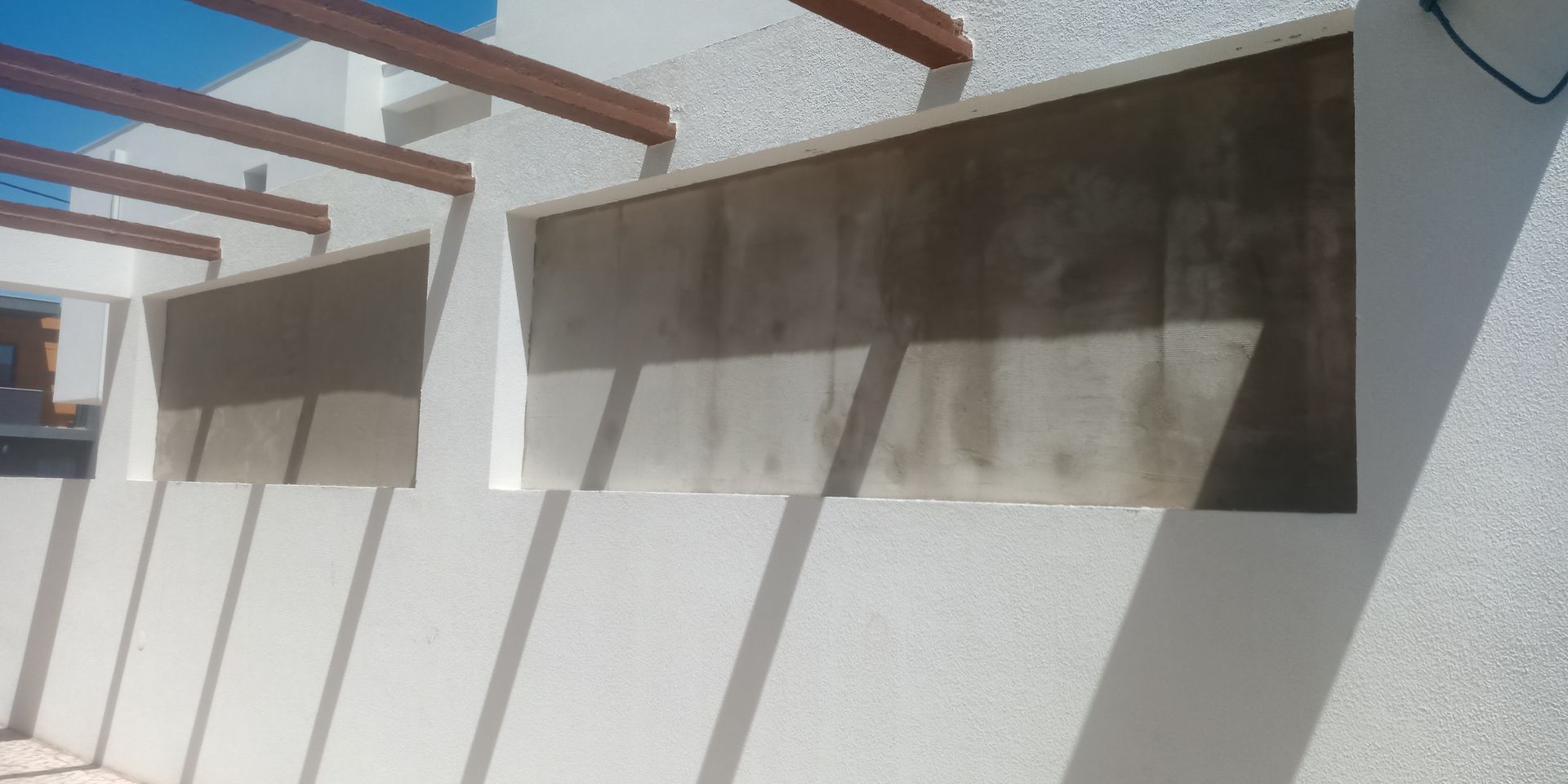 Parede externa com brises de concreto sob uma pérgola de madeira, projetando sombras.