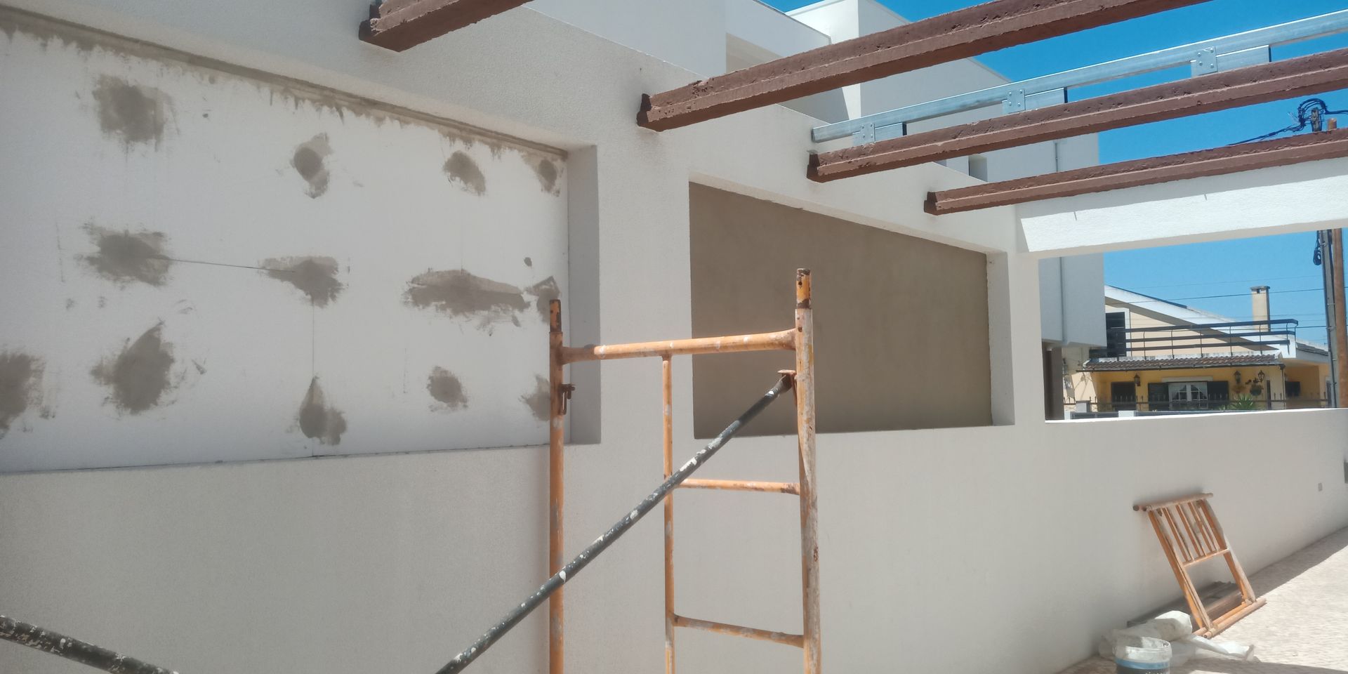 Canteiro de obras com andaimes encostados em uma parede branca; pérgola marrom acima.