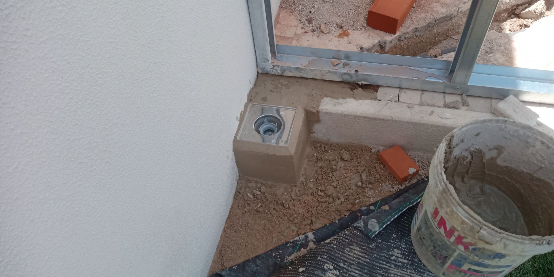 Canteiro de obras: estrutura metálica, tijolos, caçamba de concreto e parede. O solo está coberto de areia.