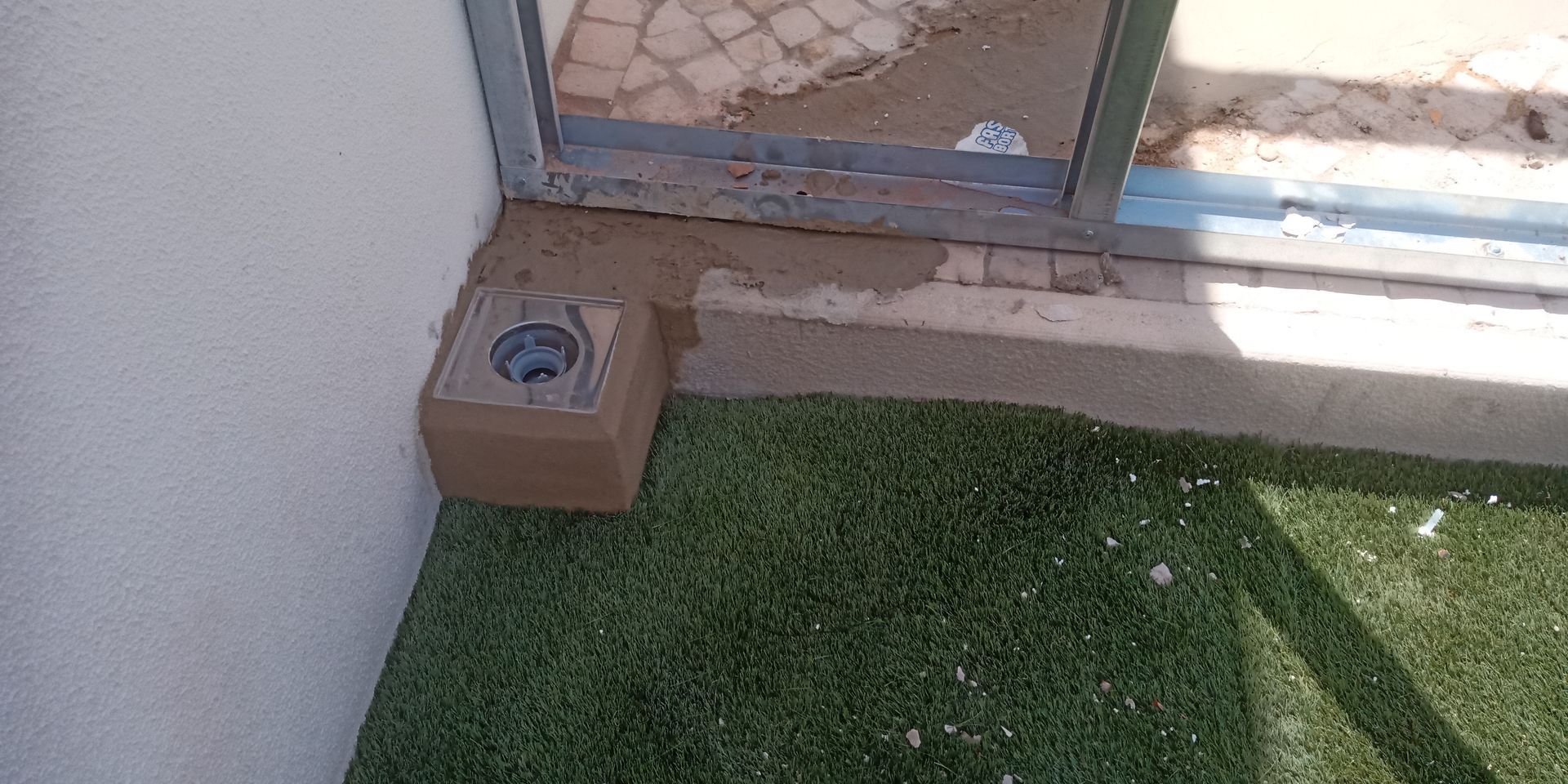Esquina de um prédio com grama artificial e um objeto quadrado marrom com um buraco no meio.