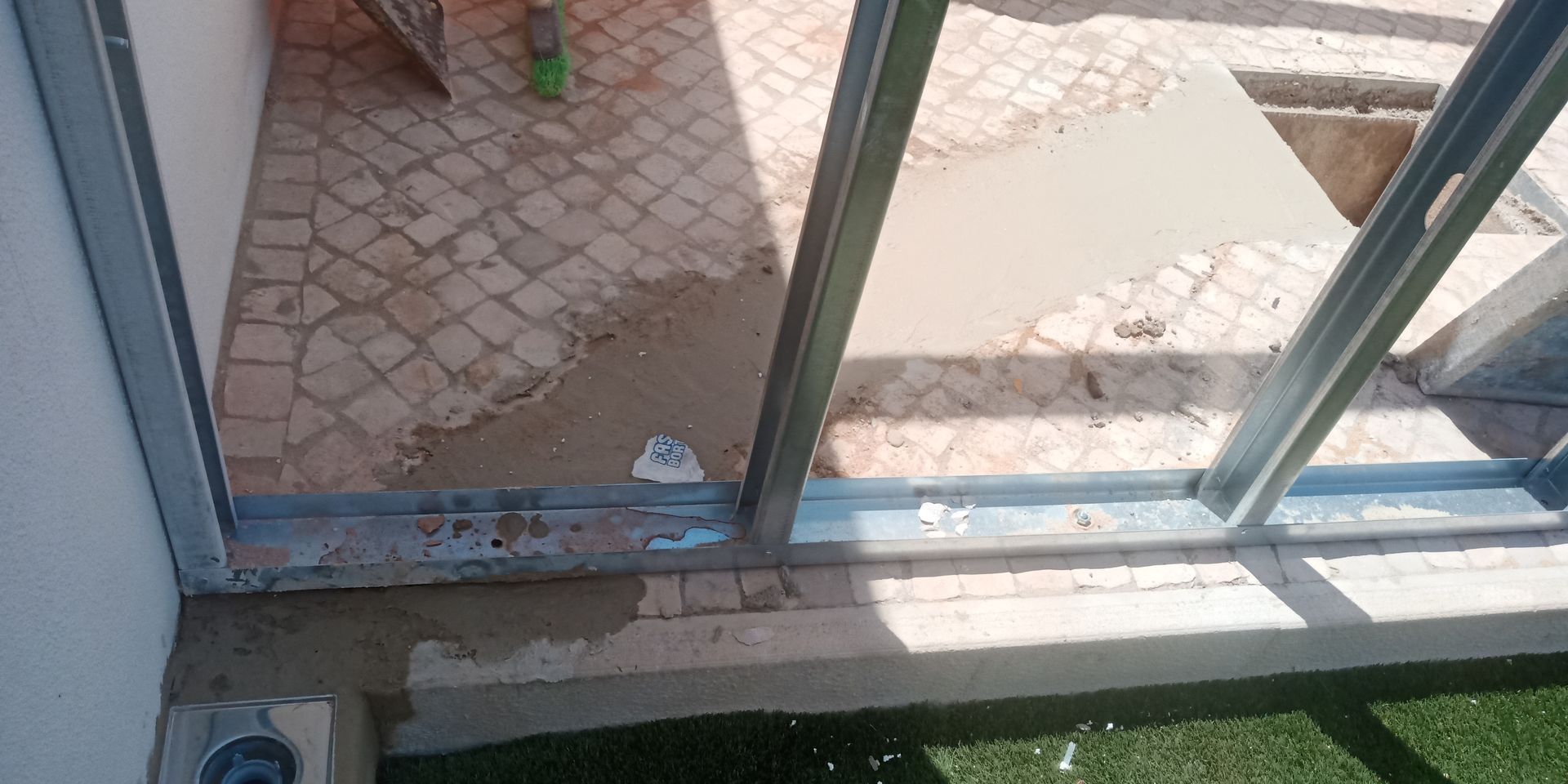 Estrutura metálica de uma porta ou janela de correr em um pátio com piso de azulejo, alvenaria e grama verde.