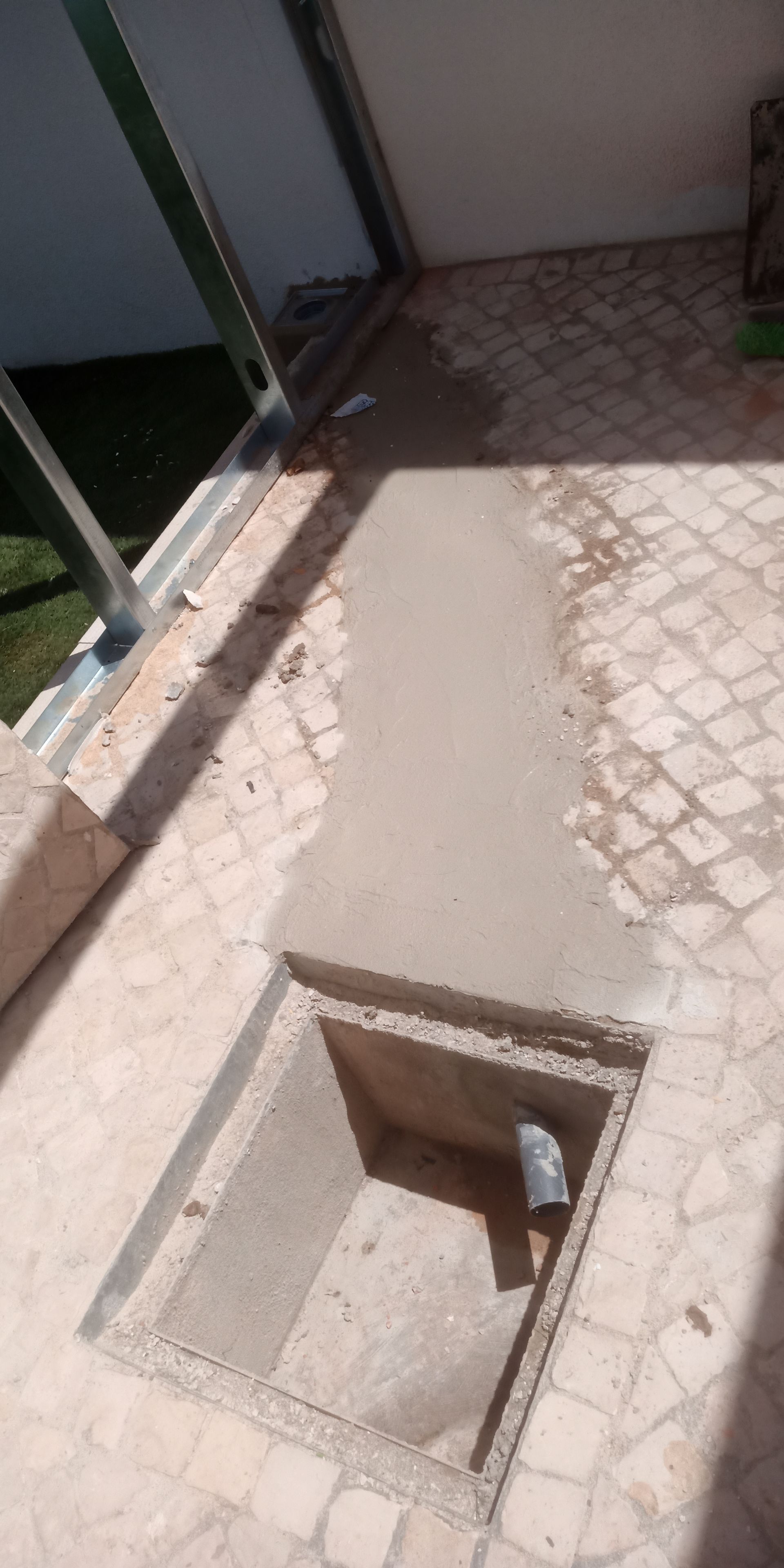 Abertura quadrada em um piso de pátio revestido com azulejos e com um remendo de concreto, possivelmente para acesso ou drenagem.