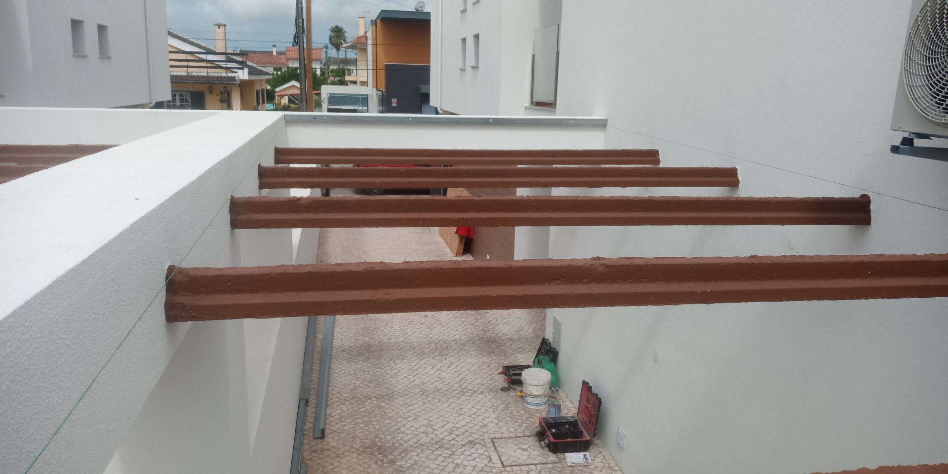 Vigas marrons se estendem sobre uma parede branca, proporcionando sombra em um espaço ao ar livre.