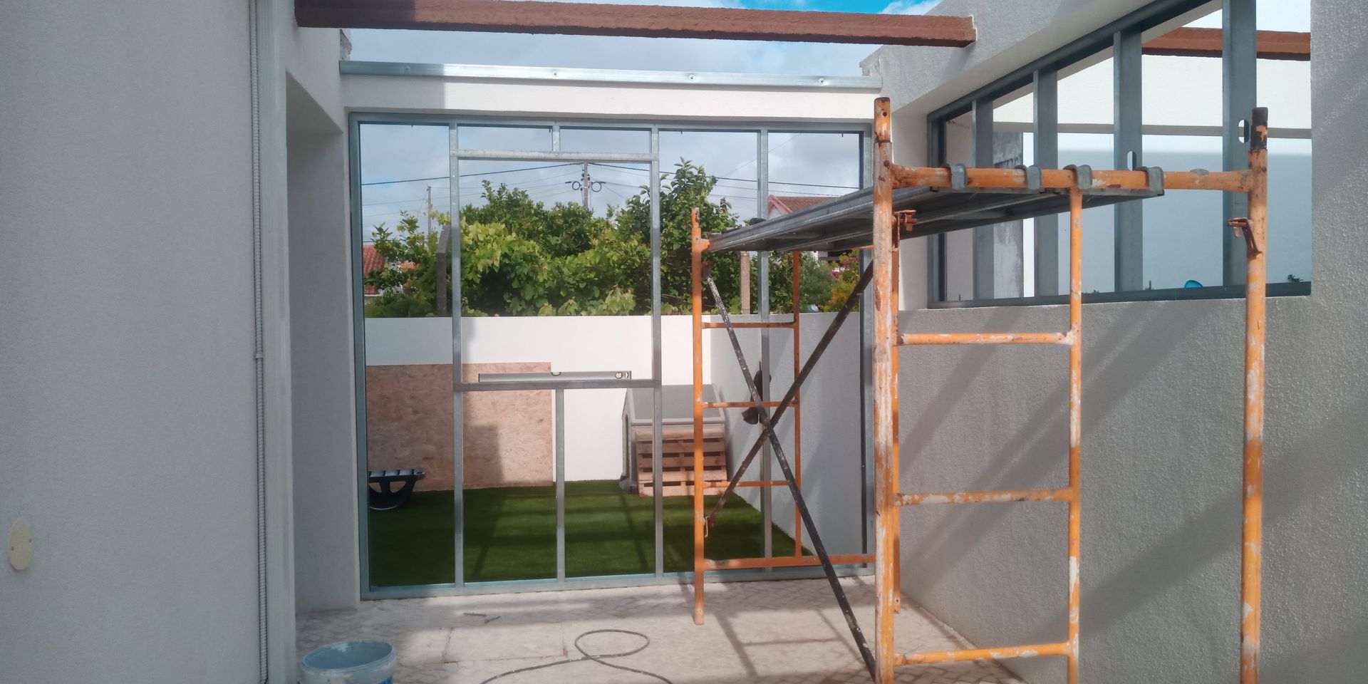 Vista de um pátio com porta de vidro e moldura de janela com andaimes. Gramado verde e árvores visíveis através da porta.