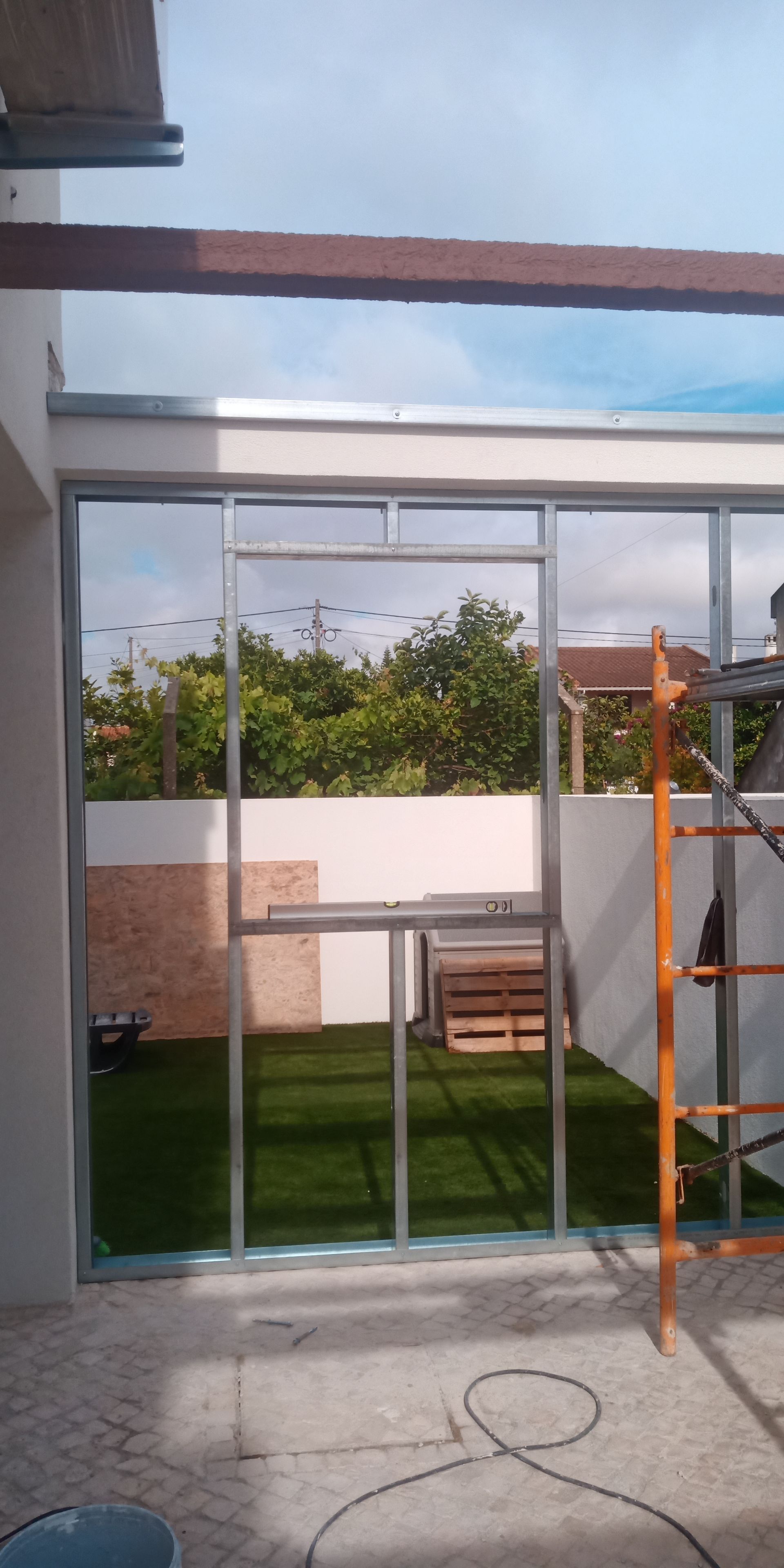 Vista exterior através de janelas de vidro inacabadas; gramado verde, parede branca, céu azul. Andaimes visíveis.