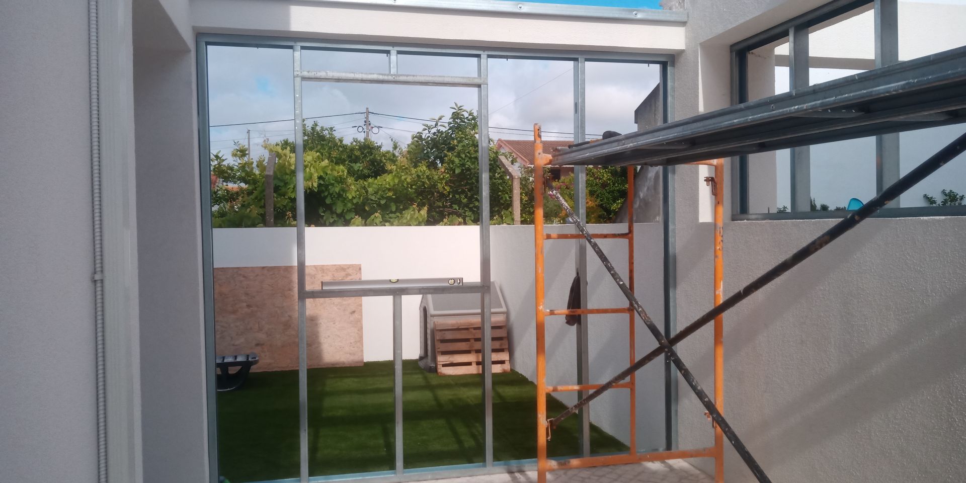 Vista interior, porta de vidro com andaime. Através da porta, avista-se um pequeno jardim com algumas árvores.