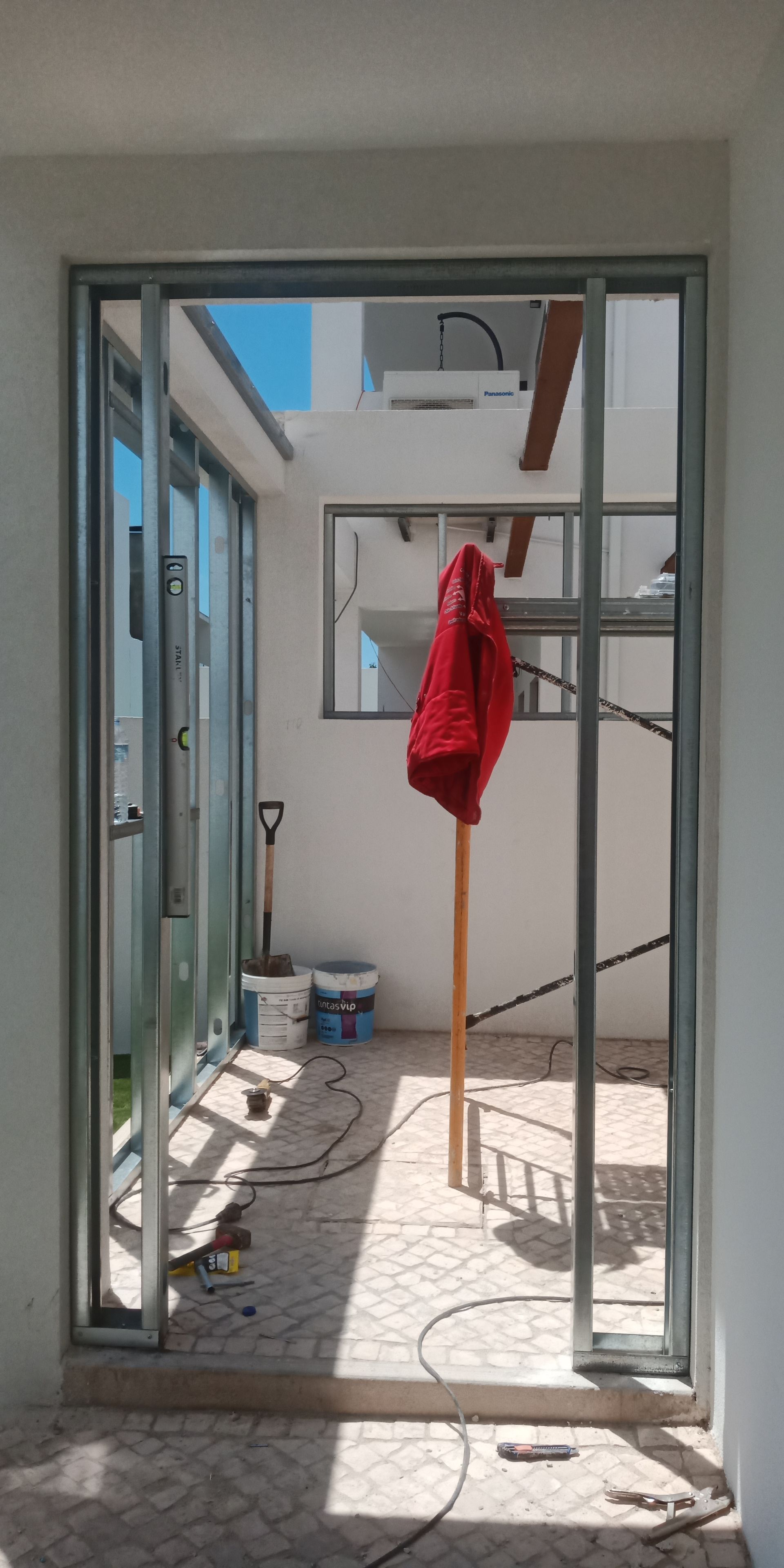 Porta com estrutura metálica e uma jaqueta vermelha pendurada do lado de dentro, revelando uma área externa inacabada.