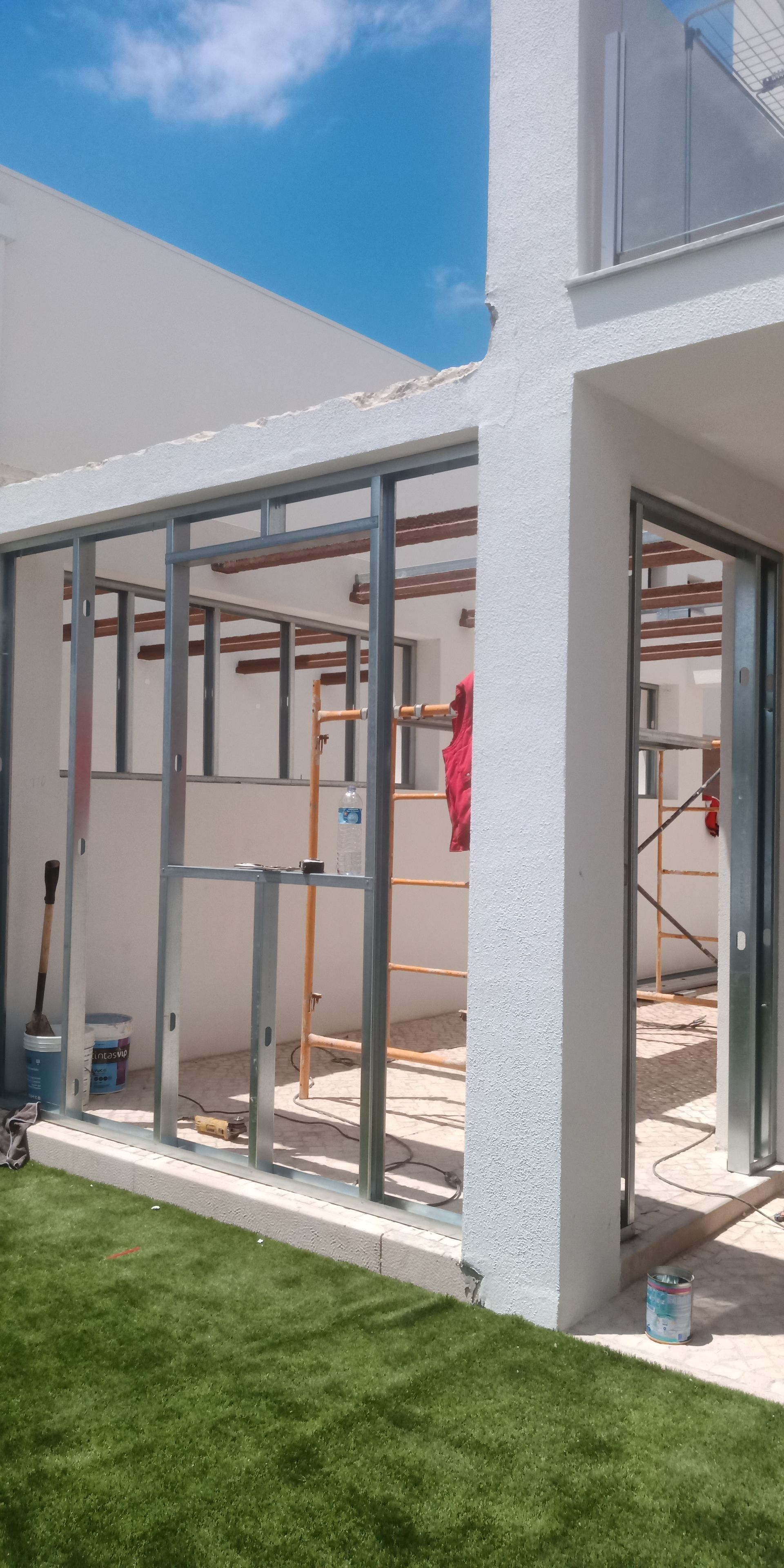 Canteiro de obras: estrutura metálica de uma extensão de edifício, escada e vegetação com céu azul.