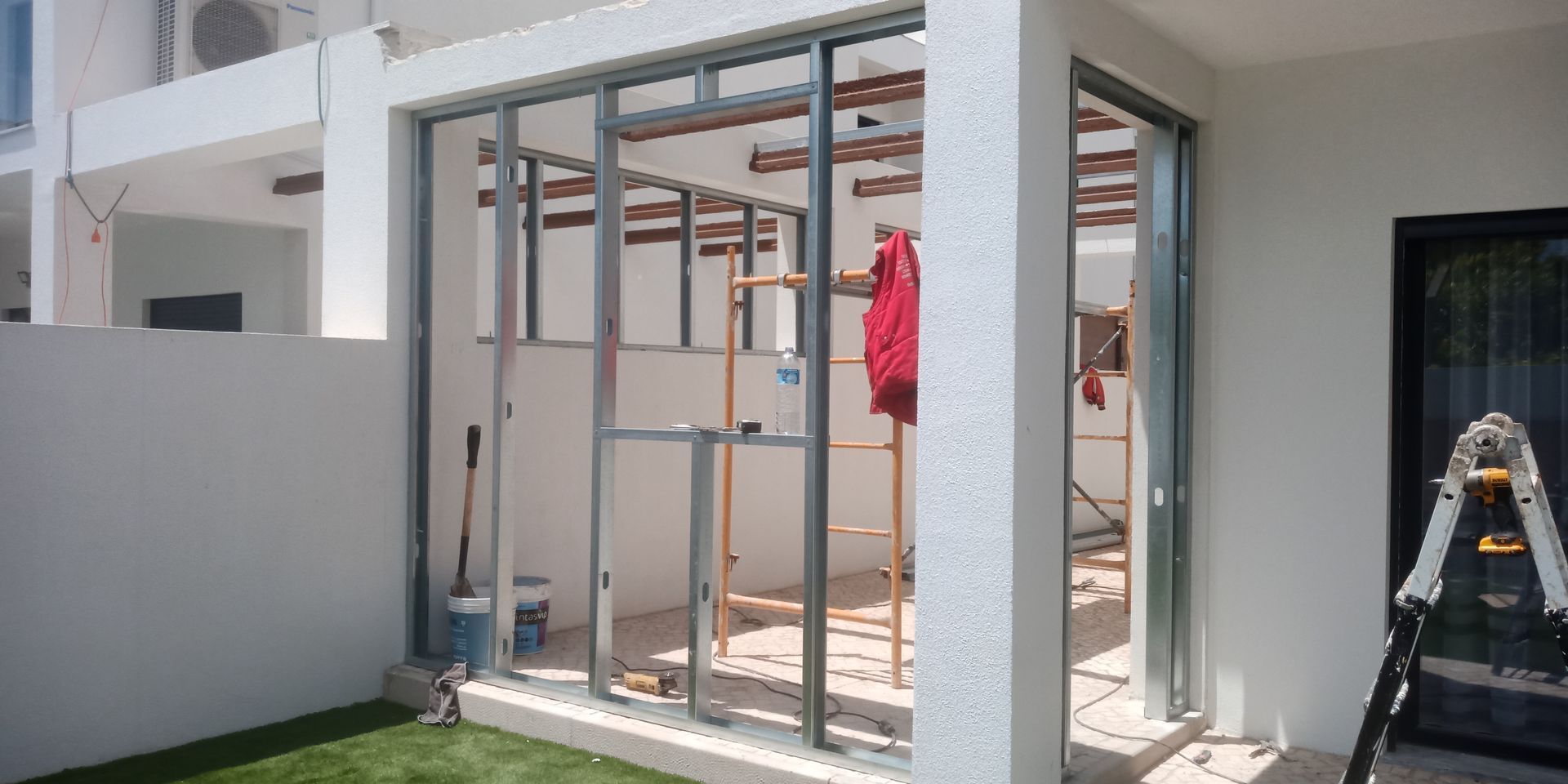 Construção de um cômodo externo com estrutura metálica. Uma peça de roupa vermelha está pendurada no interior. Paredes brancas, grama e uma escada são visíveis.
