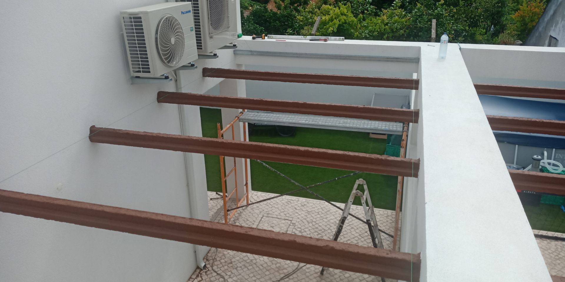 Aparelhos de ar condicionado e vigas de madeira em um telhado branco, com vista para um quintal verde e árvores.