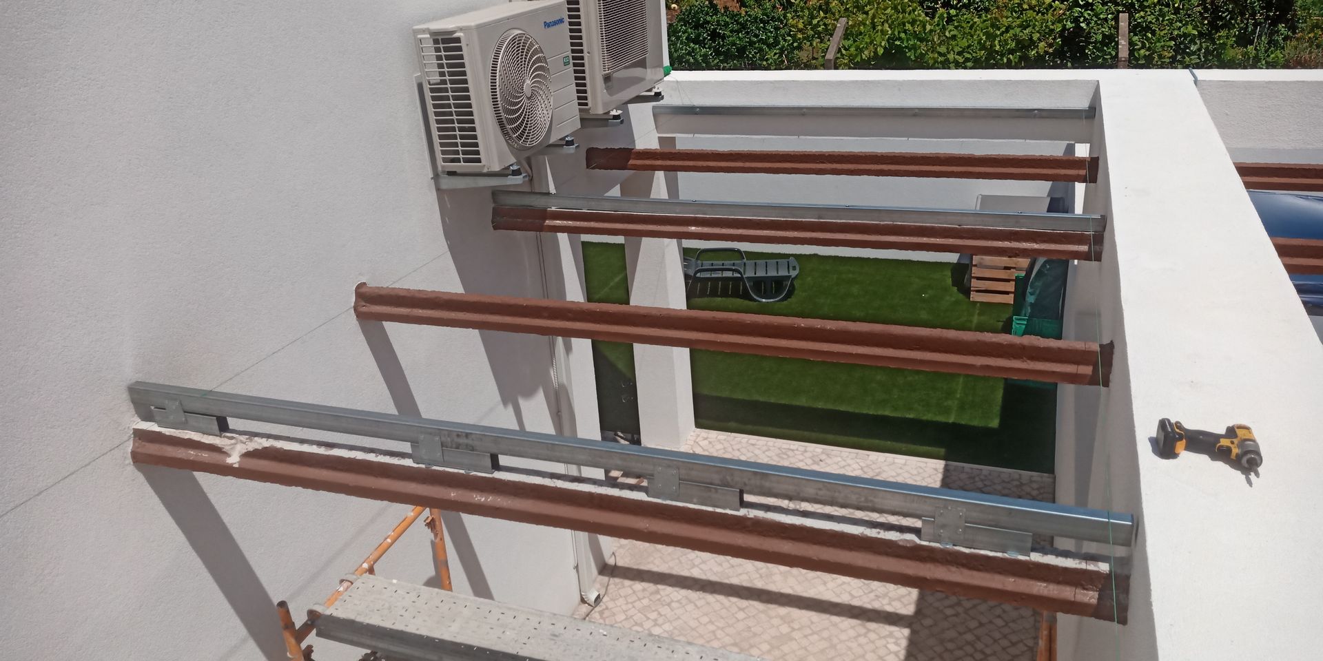 Construção de uma pérgola com vigas cor de cobre em uma parede branca; unidade de ar condicionado no canto superior esquerdo.