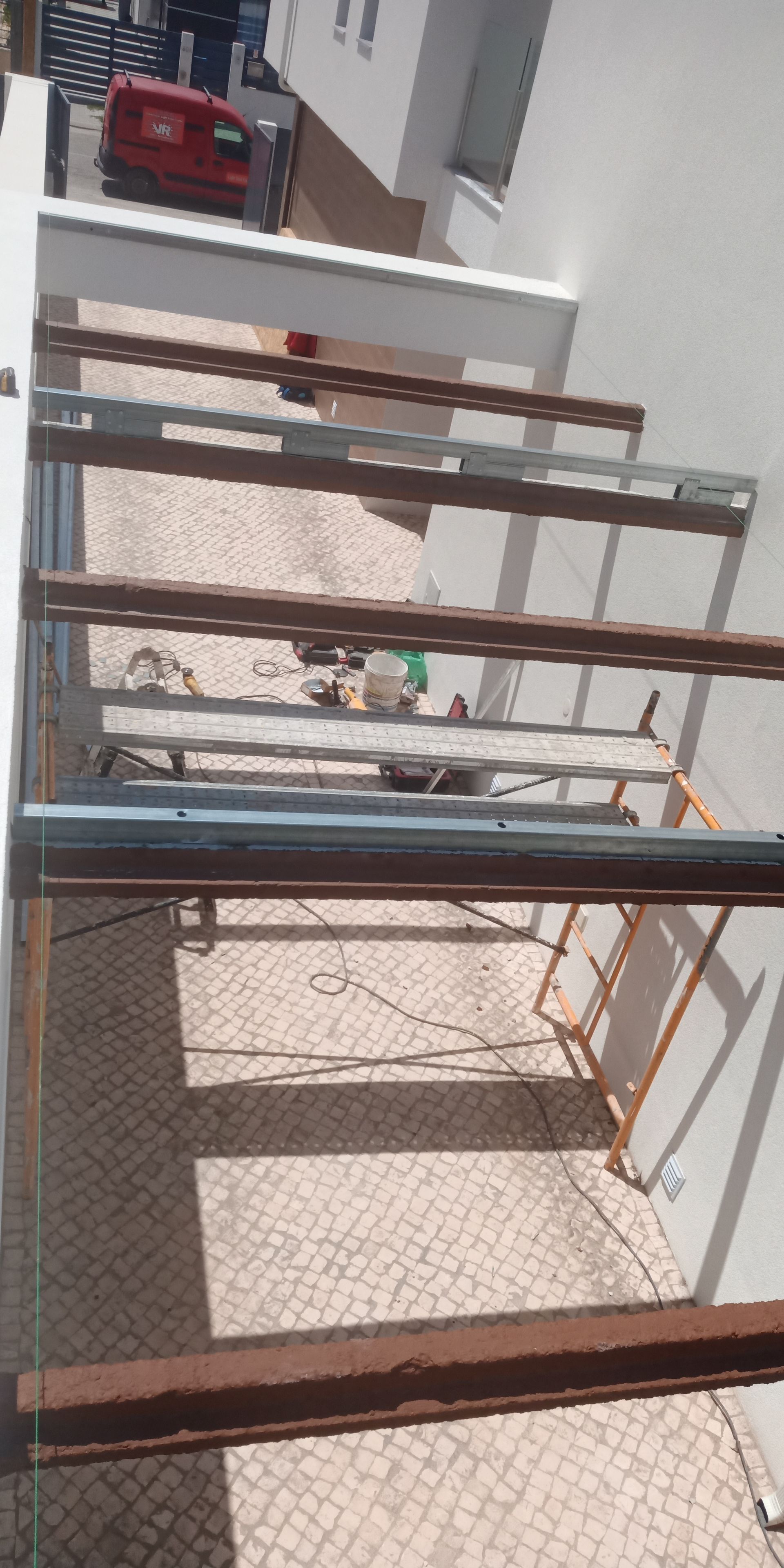 Estrutura metálica com um veículo vermelho estacionado na rua. Área sombreada com piso de paralelepípedos.