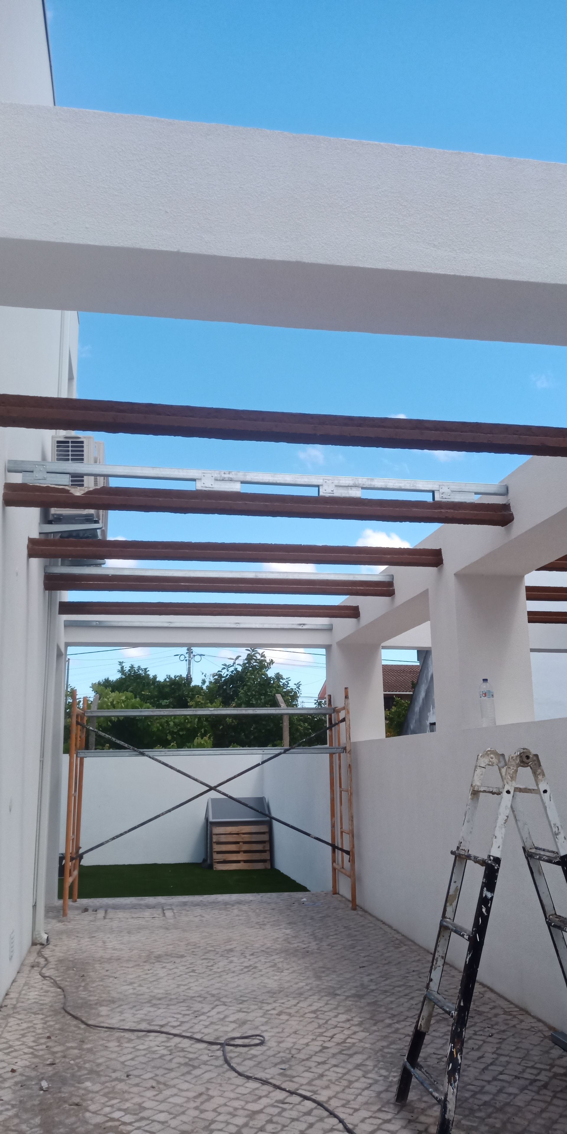 Uma garagem com paredes brancas, vigas de madeira marrom e um céu azul ao fundo. Uma escada está encostada na parede.