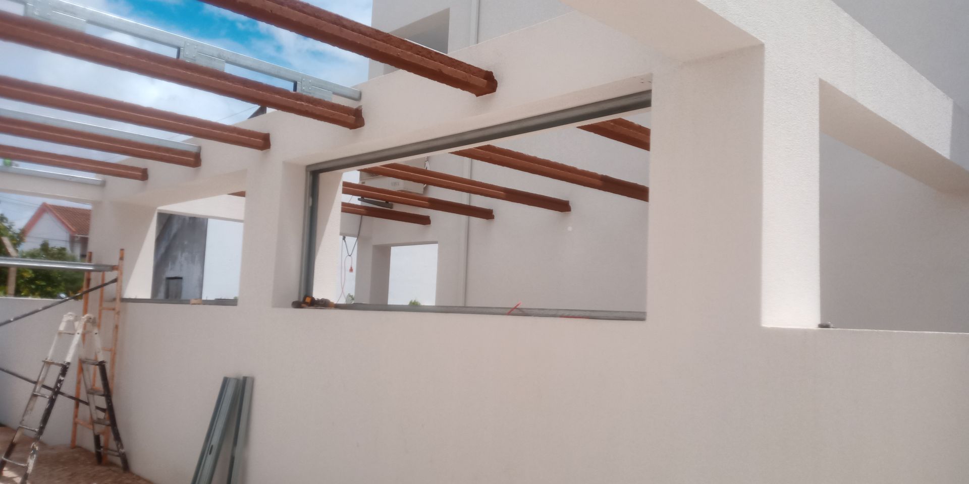 Canteiro de obras: vigas marrons emolduram uma abertura, paredes brancas, escadas e um céu azul ao fundo.
