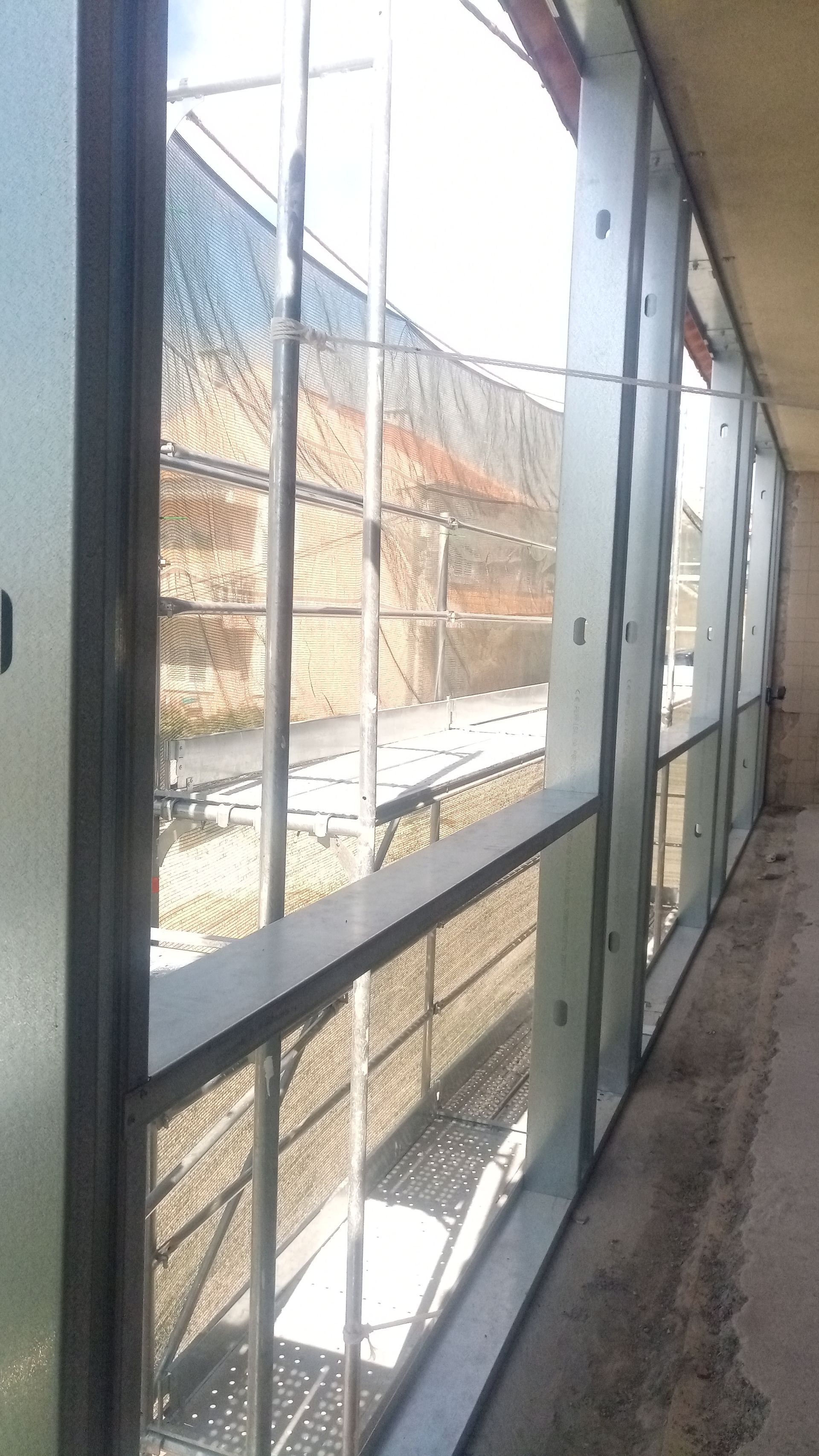 Vista exterior através de uma parede envidraçada. Estruturas metálicas sustentam os painéis de vidro; um caminho e uma construção são visíveis do lado de fora.