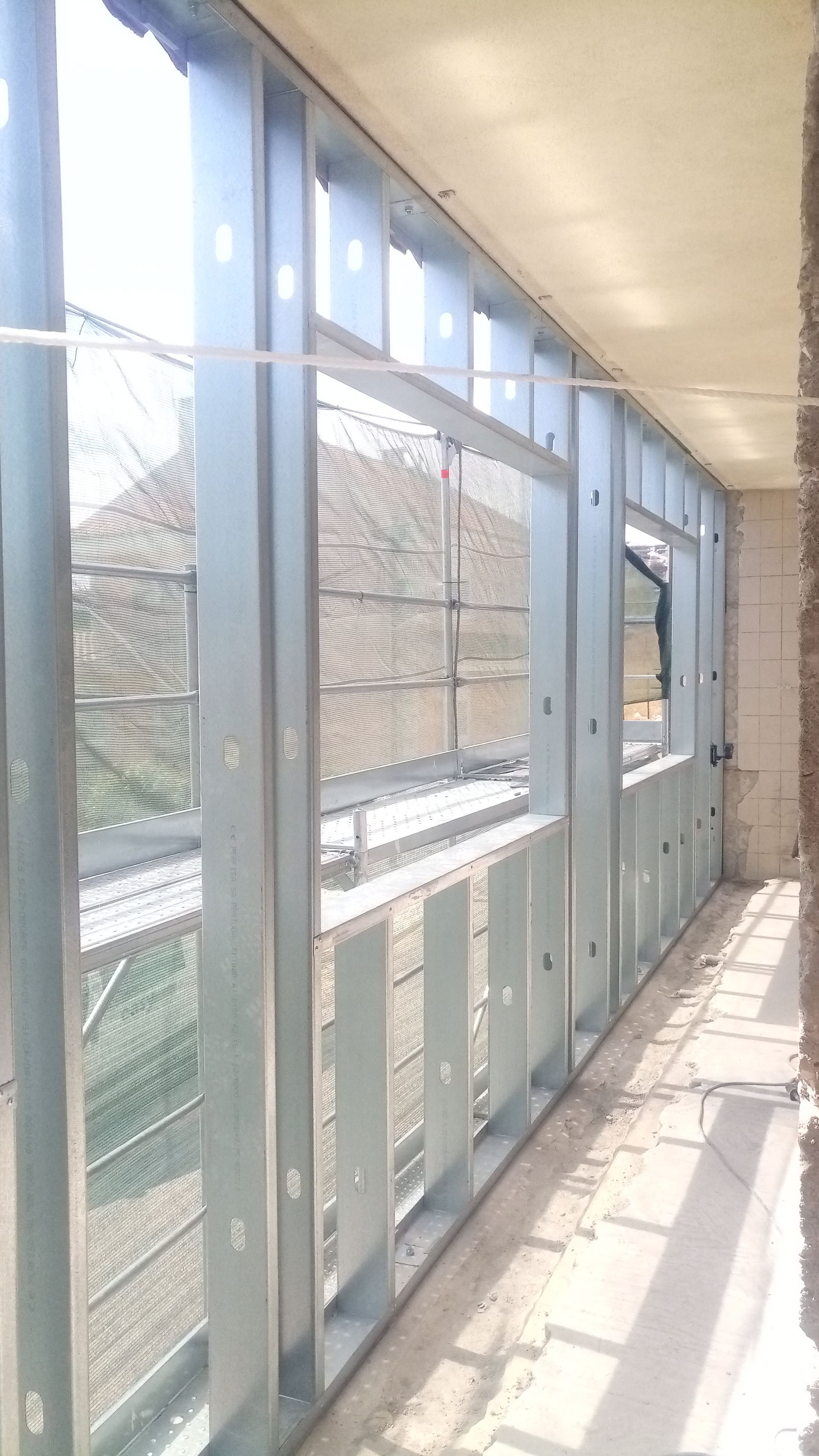 Estrutura metálica para janela e parede, dentro de um prédio em construção, com vista para um pátio.