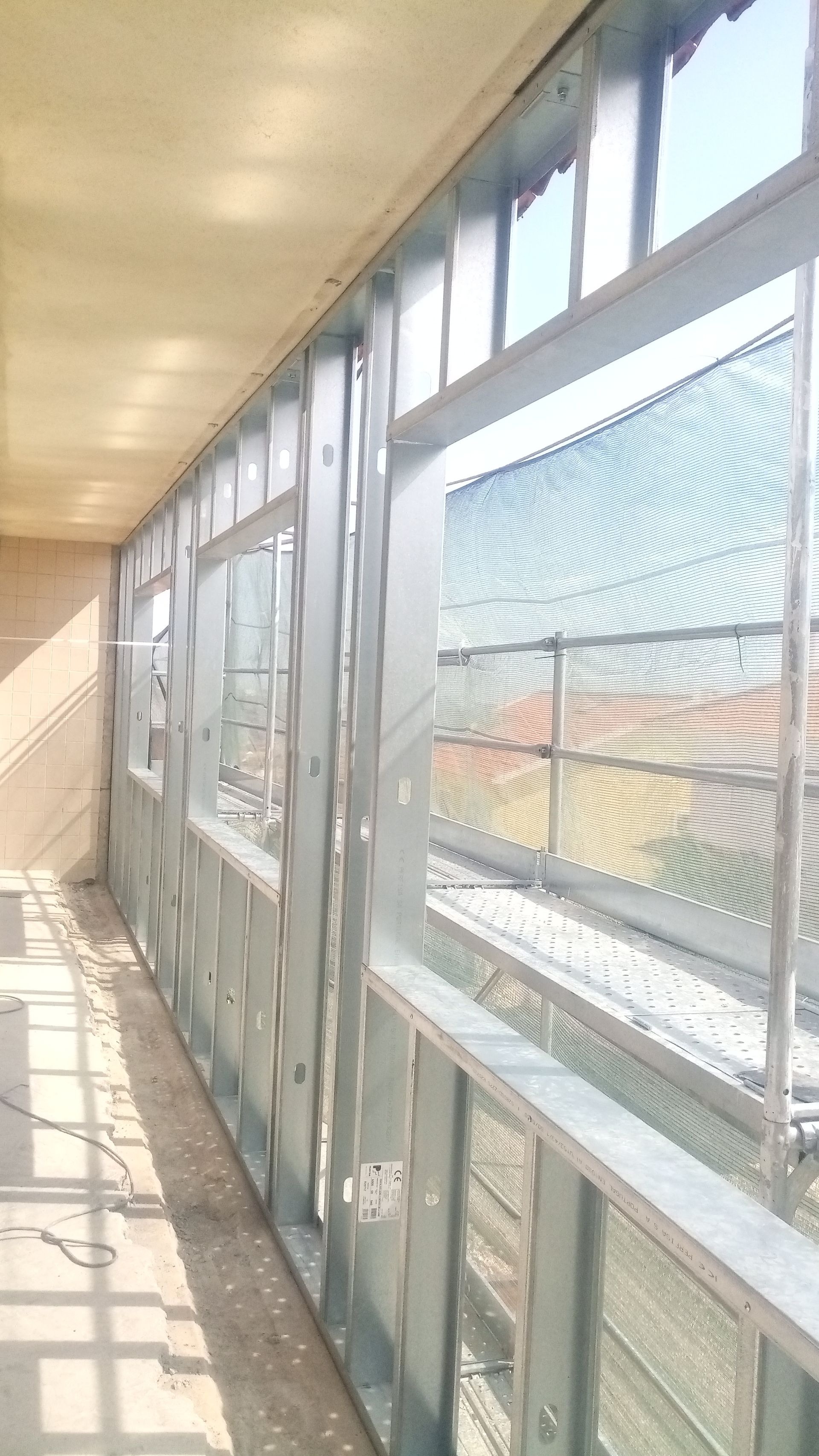 Vista interna de um edifício em construção, mostrando a estrutura de aço ao redor das janelas e o teto inacabado.