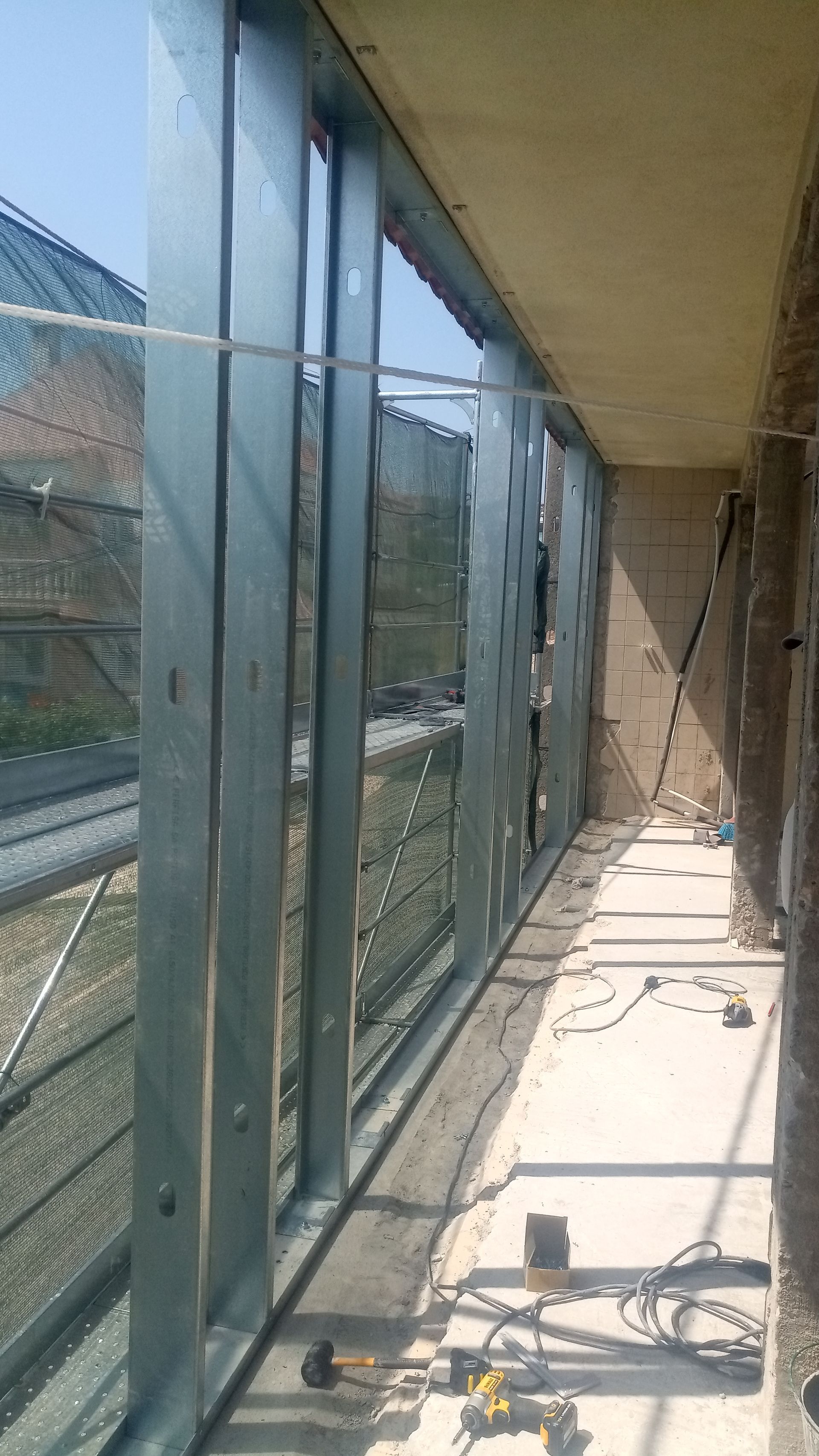 Estrutura metálica da parede externa de um edifício. Uma tela de malha é visível atrás da estrutura.