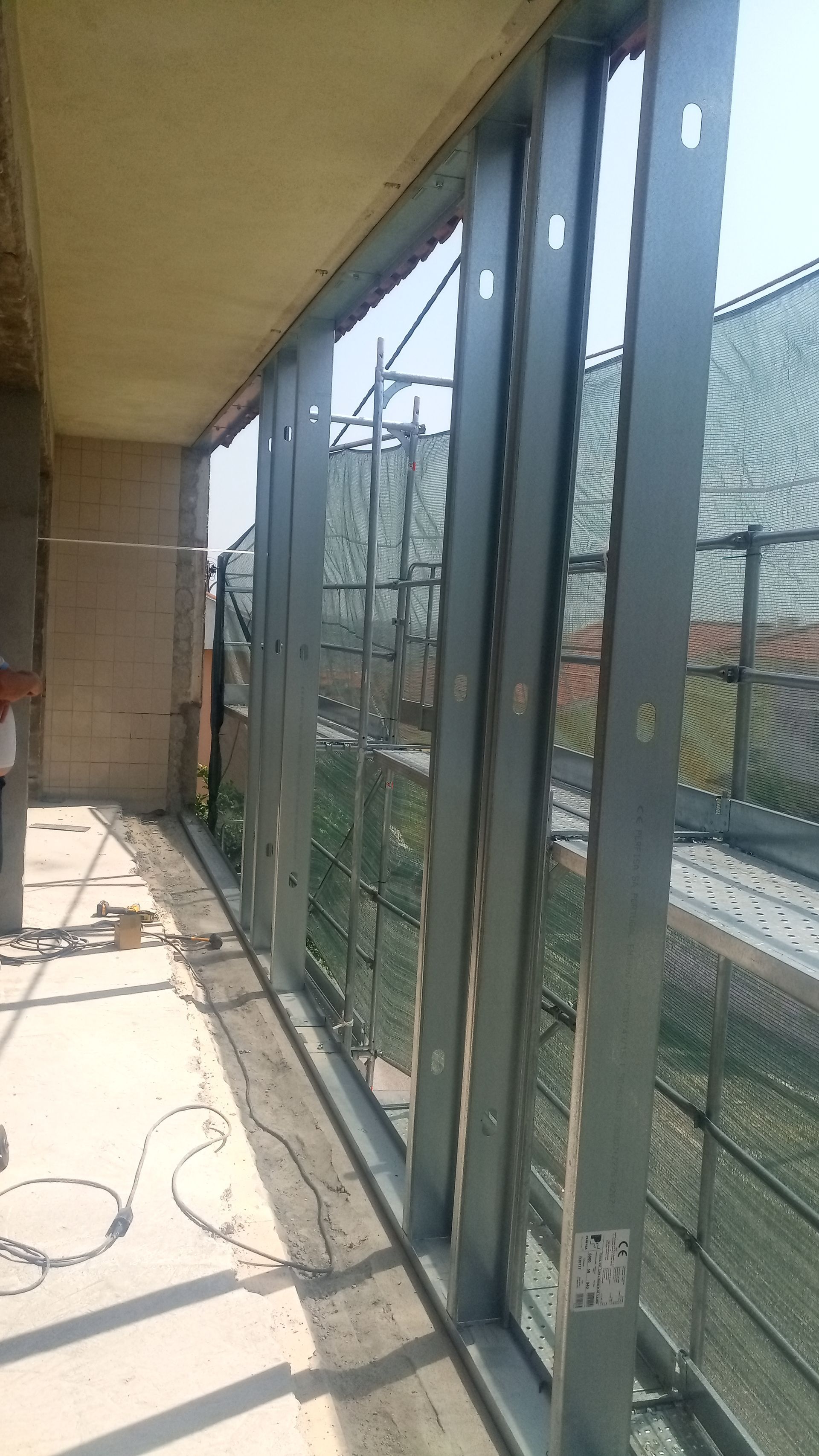 Estrutura de aço para uma varanda, com parede de concreto e guarda-corpo de tela metálica ao fundo.