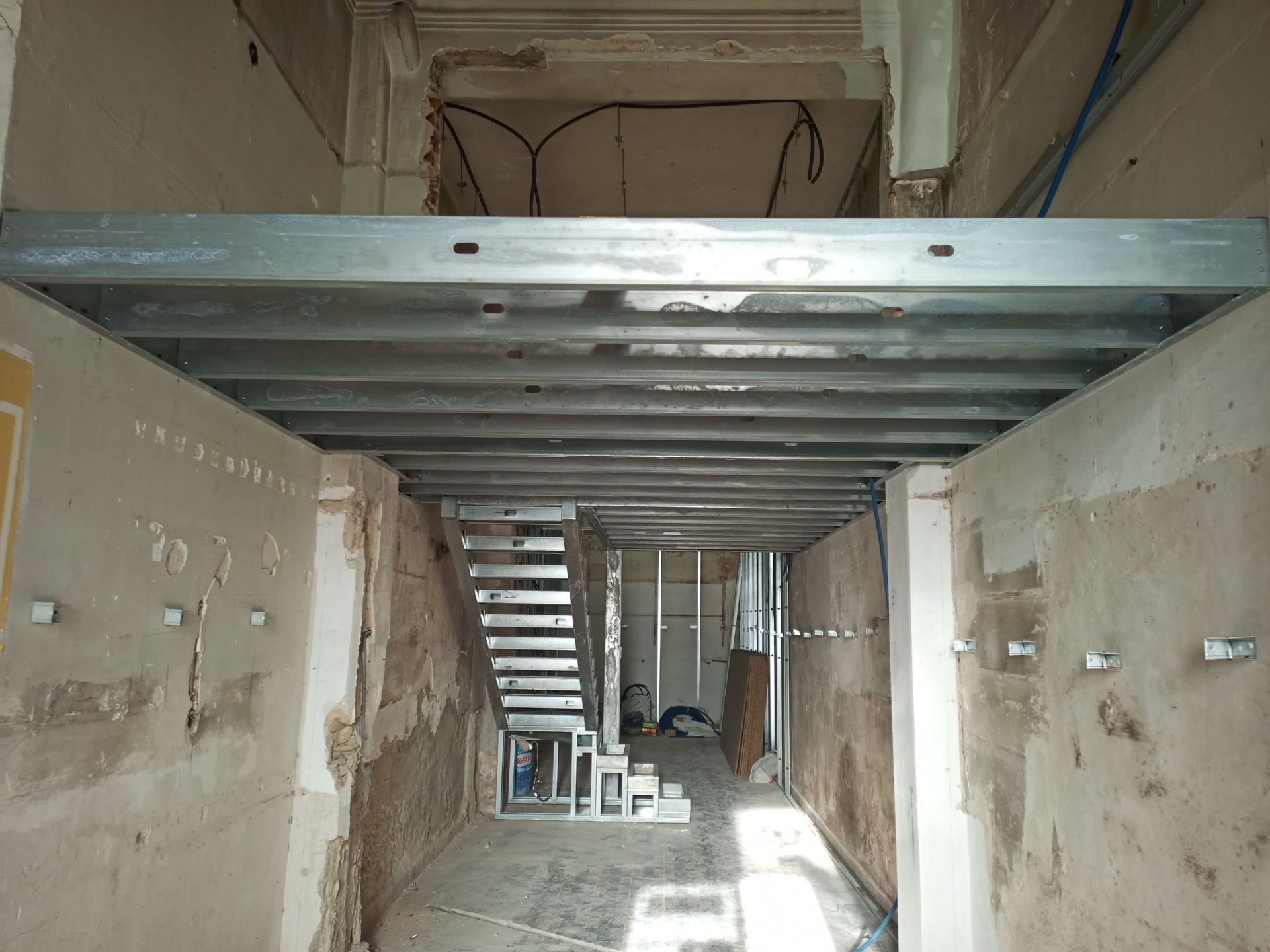 Vista interior de um edifício em renovação, com vigas metálicas e uma escadaria visíveis ao fundo.