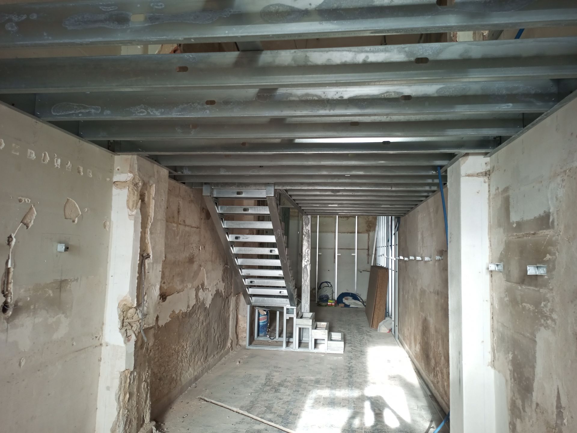 Vista interior de um edifício em construção, com vigas de aço expostas, uma escadaria e paredes inacabadas.